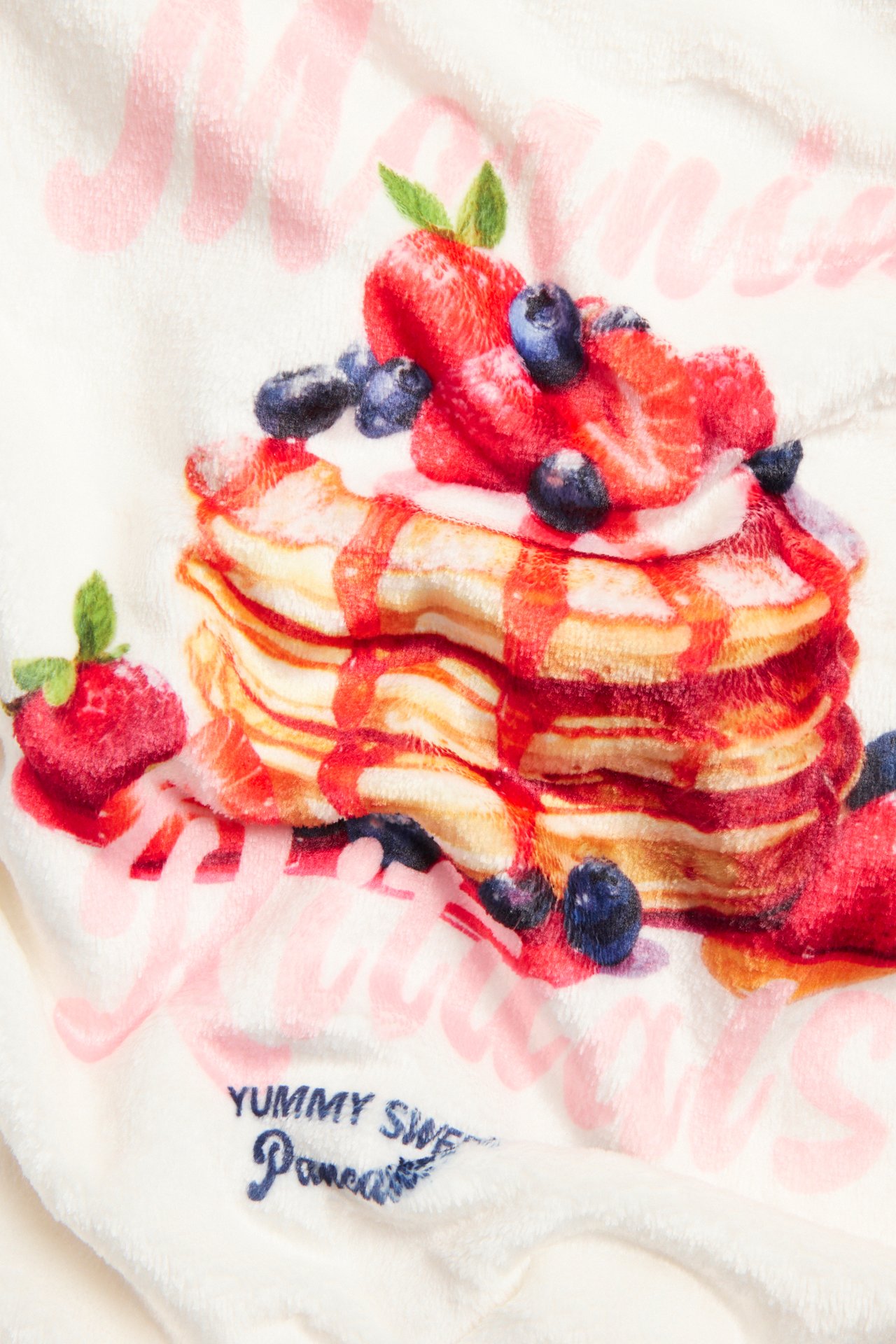 Närbild av offwhite fleecehoodie tyg med färgglatt pannkaks-, jordgubbs- och blåbärstryck samt text.