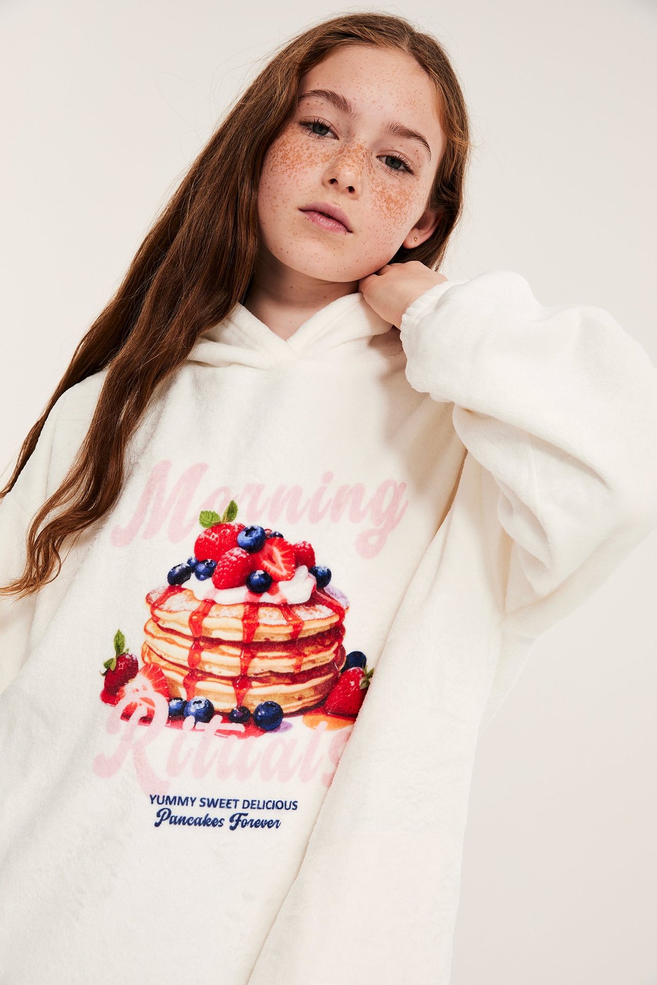 Nærbilde forfra av et barn i en offwhite fleecehoodie med fargerikt pannekake- og bærprint.