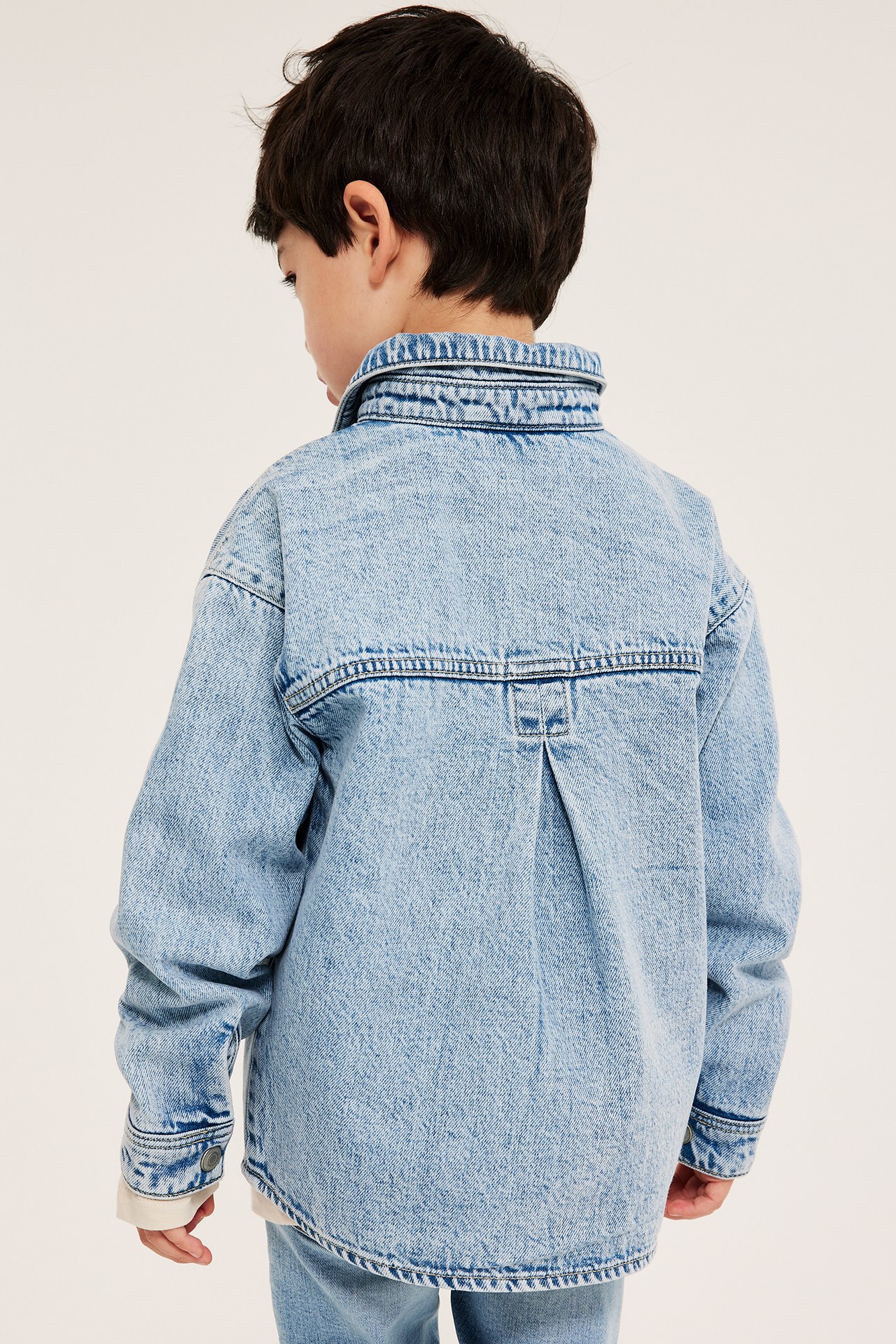 Bakfra, et barn i lys blå denim-overskjorte med avslappet passform over en hvit skjorte.