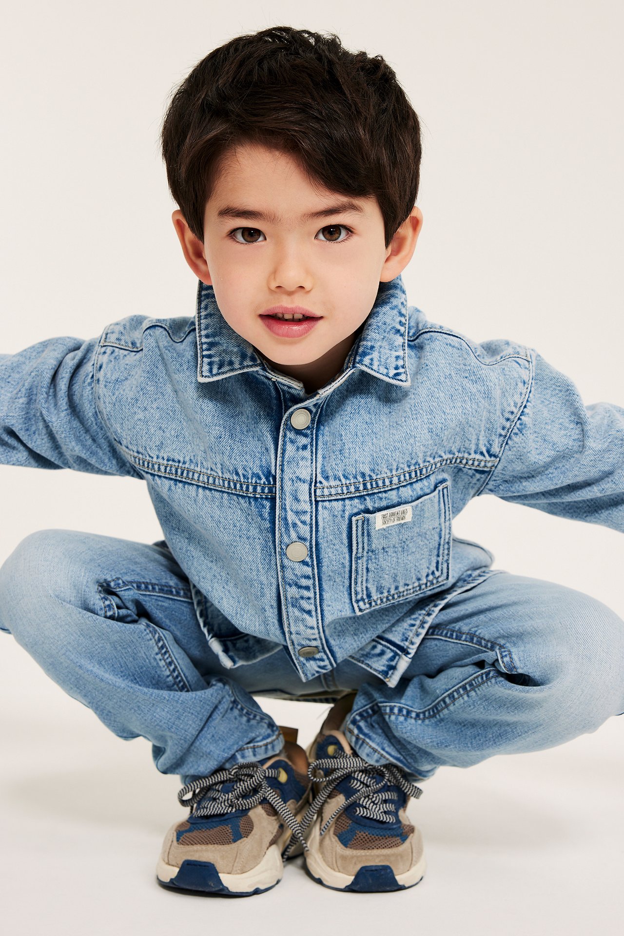 Framifrån: ett barn i en ljusblå relaxed-fit denim overshirt med tryckknappar, ljusblå jeans och flerfärgade sneakers.