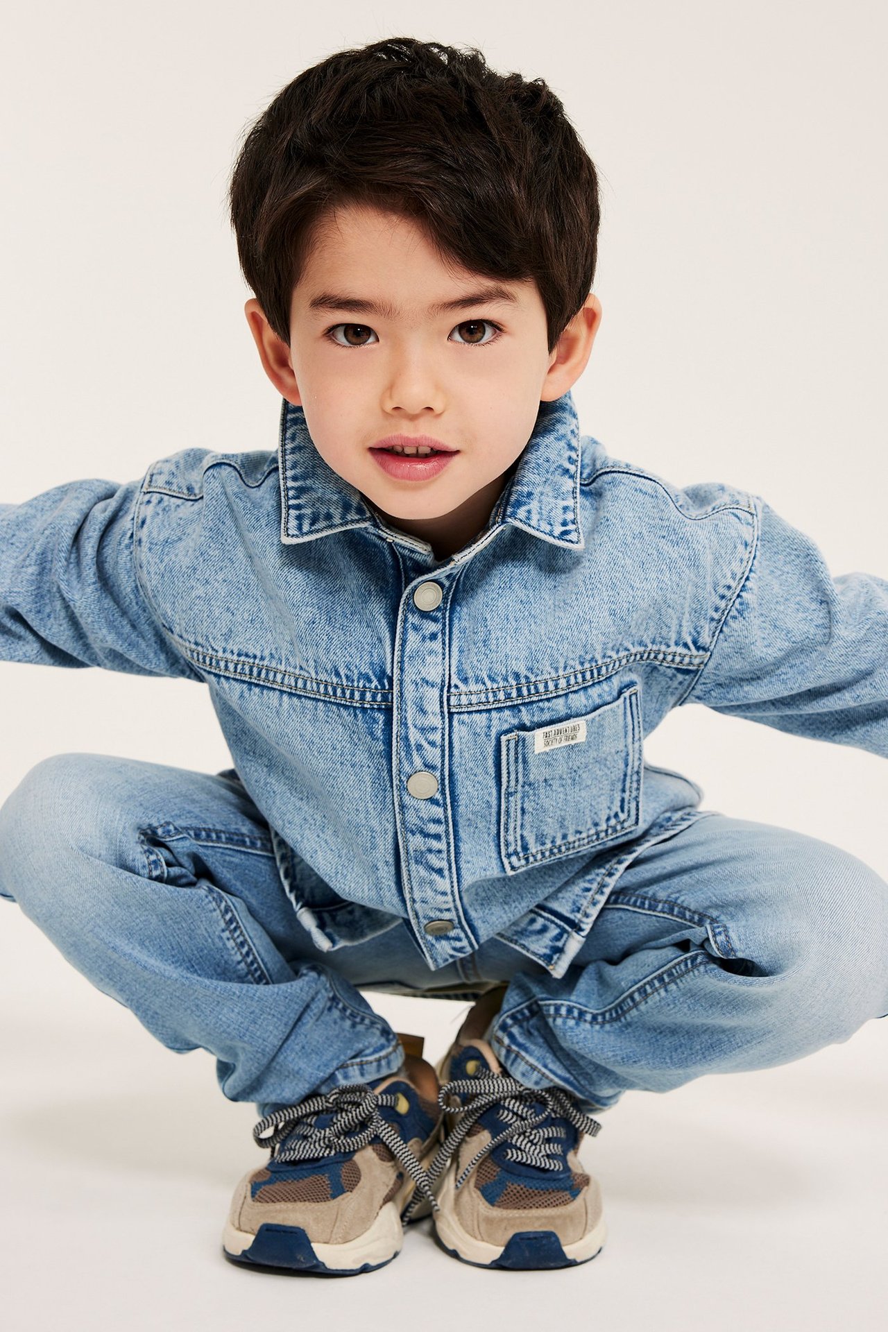 Framifrån: ett barn i en ljusblå relaxed-fit denim overshirt med tryckknappar, ljusblå jeans och flerfärgade sneakers.