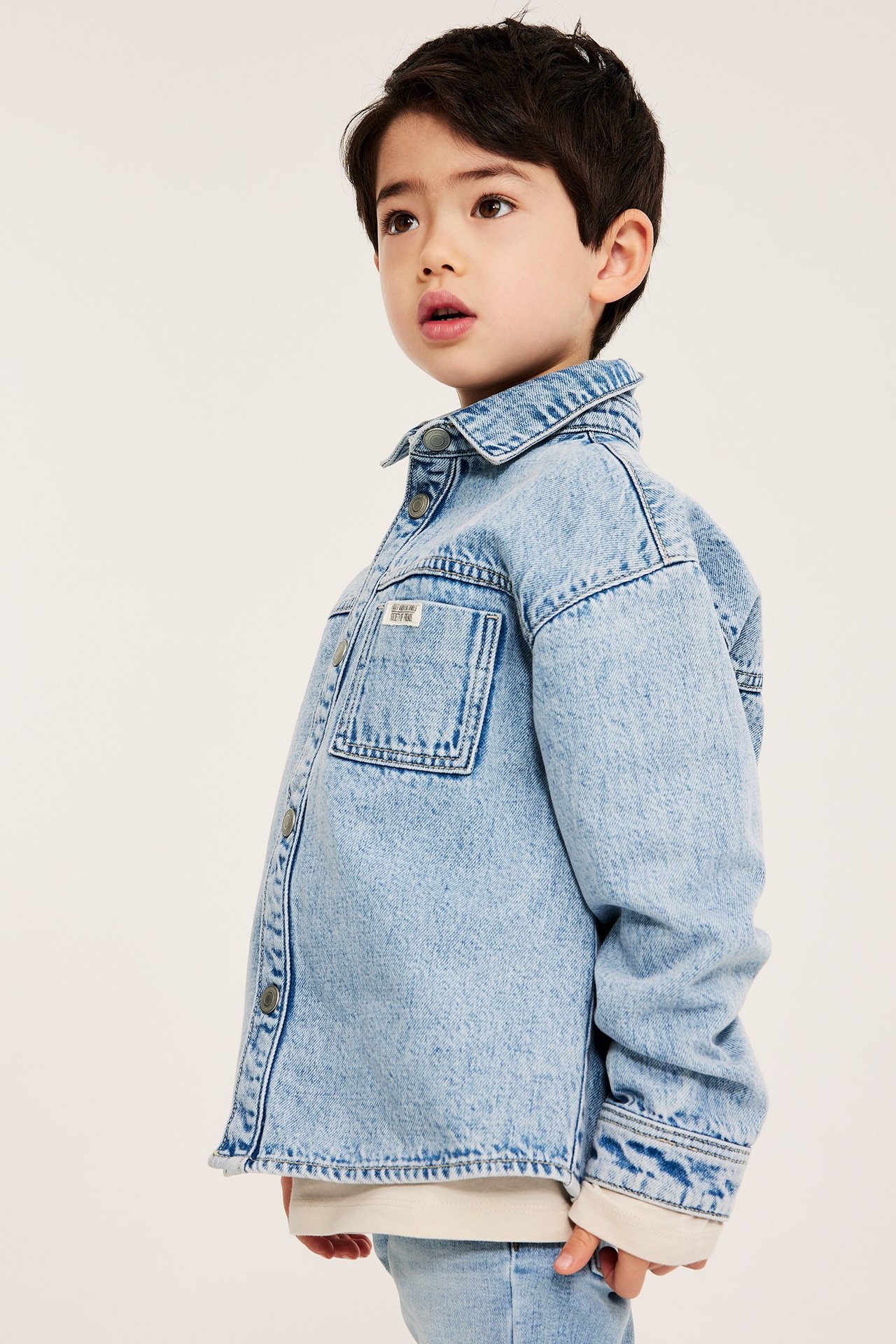 Widok z przodu: Dziecko w jasnej denimowej overshircie z zatrzaskami, na jasnej bluzce i dżinsach.