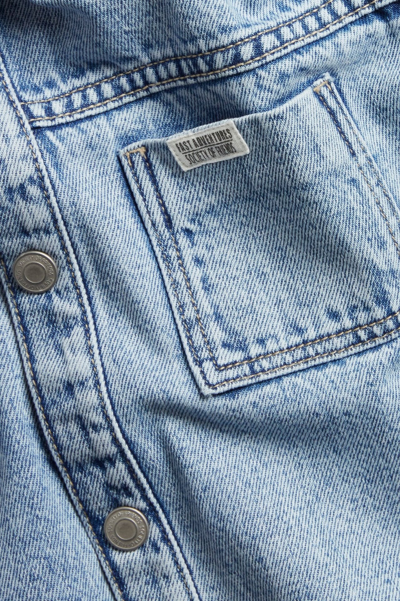 Zbliżenie na jasnoniebieską koszulę dżinsową, pokazujące kieszeń z naszywką i zatrzask.