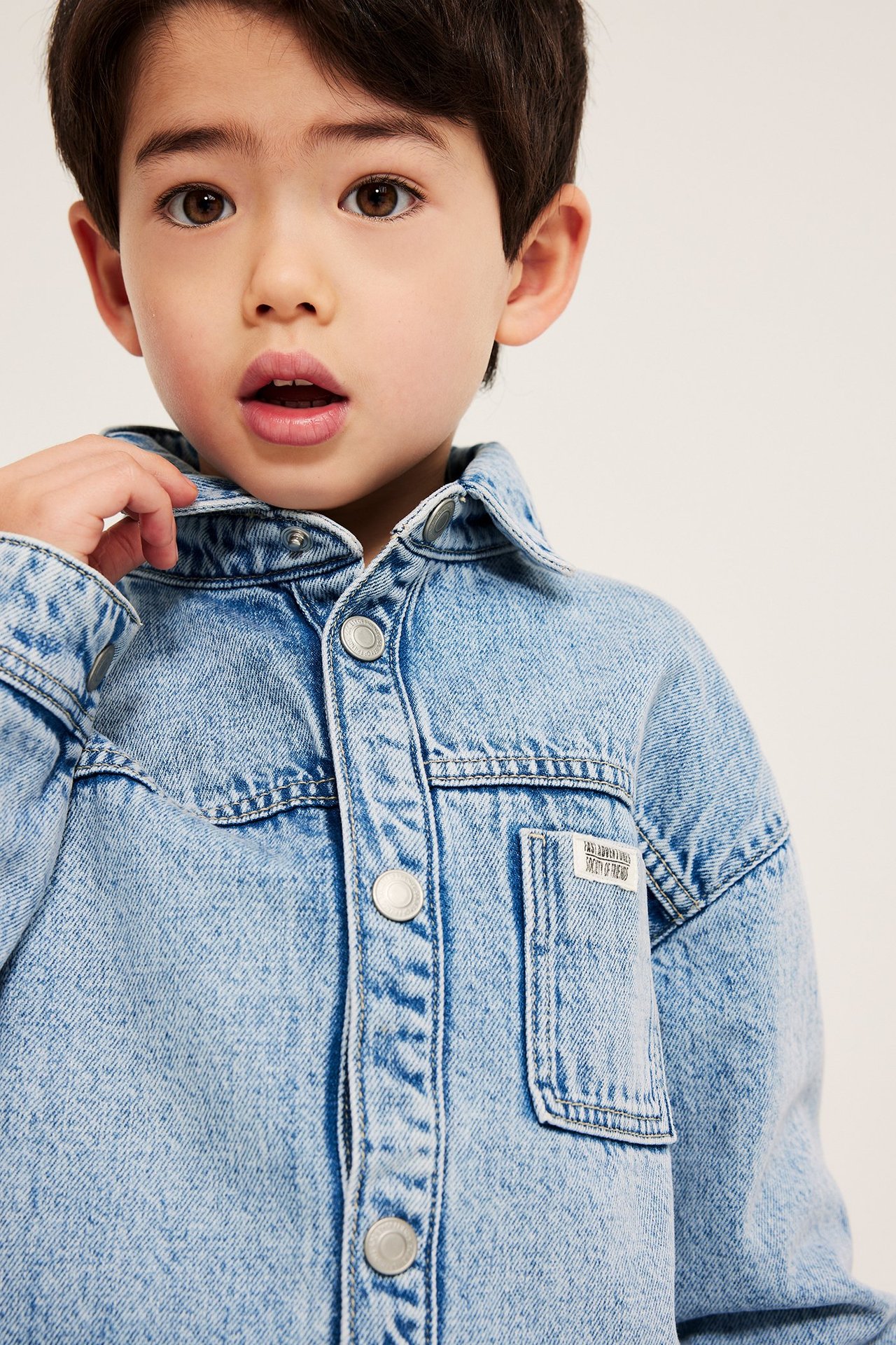 Zbliżenie dziecka w jasnej denimowej koszuli wierzchniej z zatrzaskami i kieszenią na piersi.