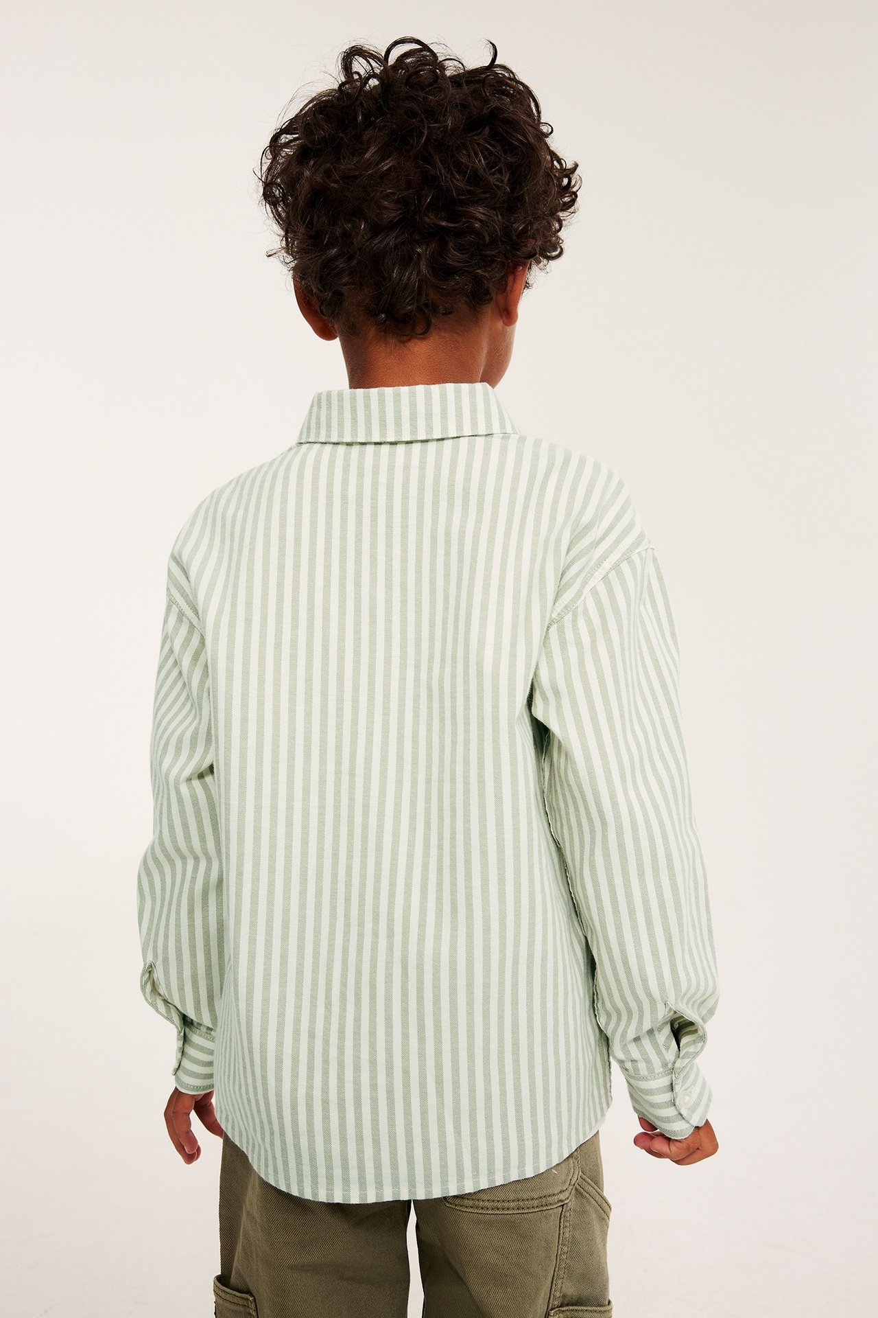 Ryggvy av ett barn i en lös ljusgrön- och vitrandig bomullsöverskjorta och khakifärgade cargobyxor.