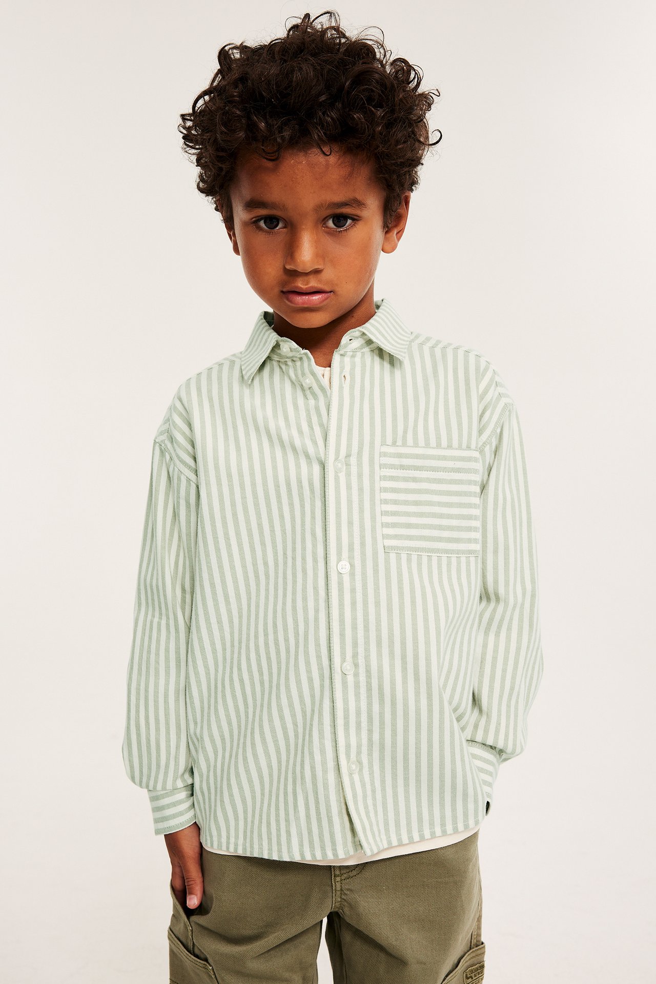 Framifrån: Pojke i ljusgrön-vit randig bomullsskjorta med ficka, och olivgröna cargobyxor.