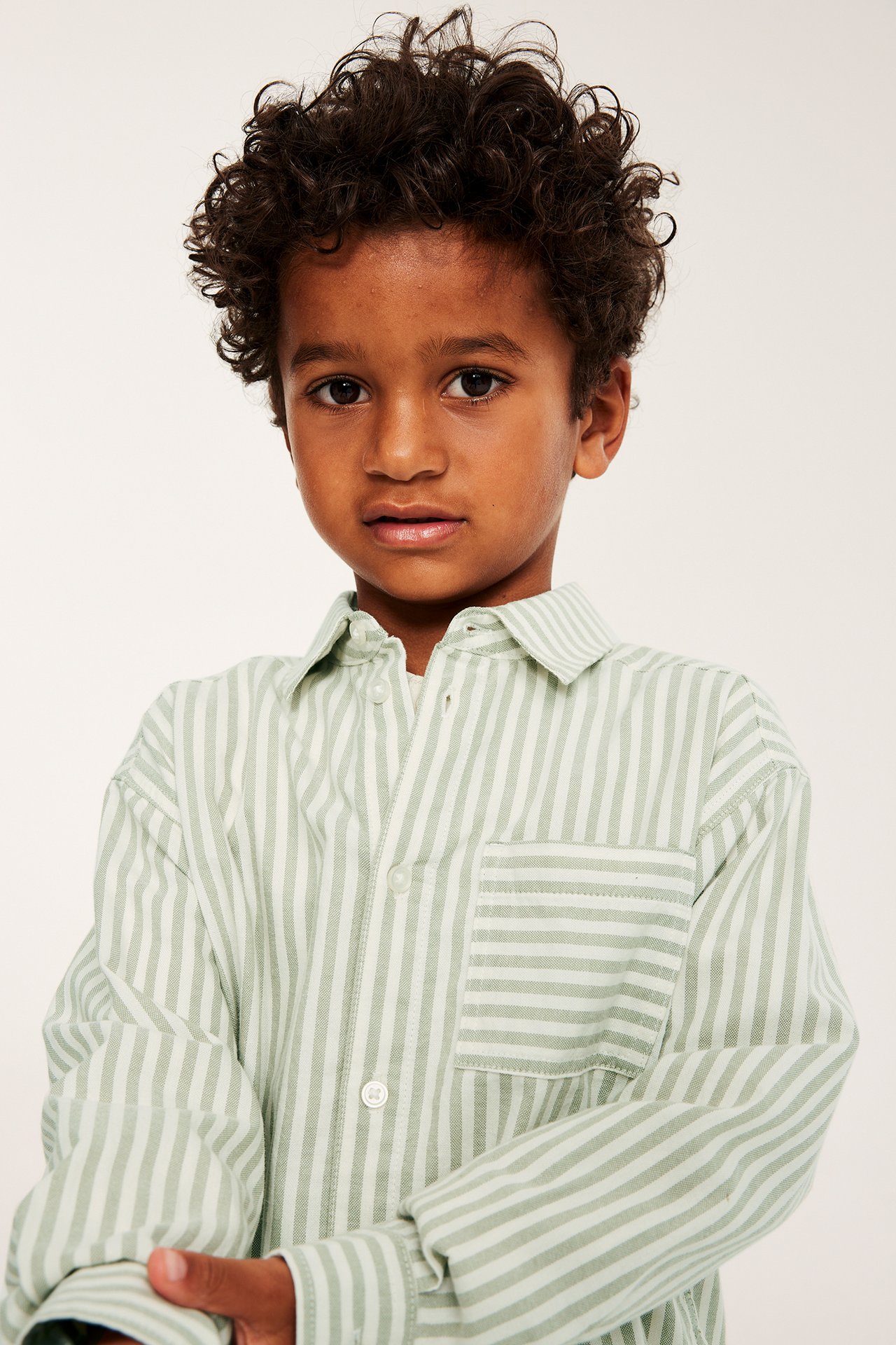 Widok z przodu, dziecko w jasnozielonej, białej bawełnianej koszuli w paski, guziki i kieszeń.