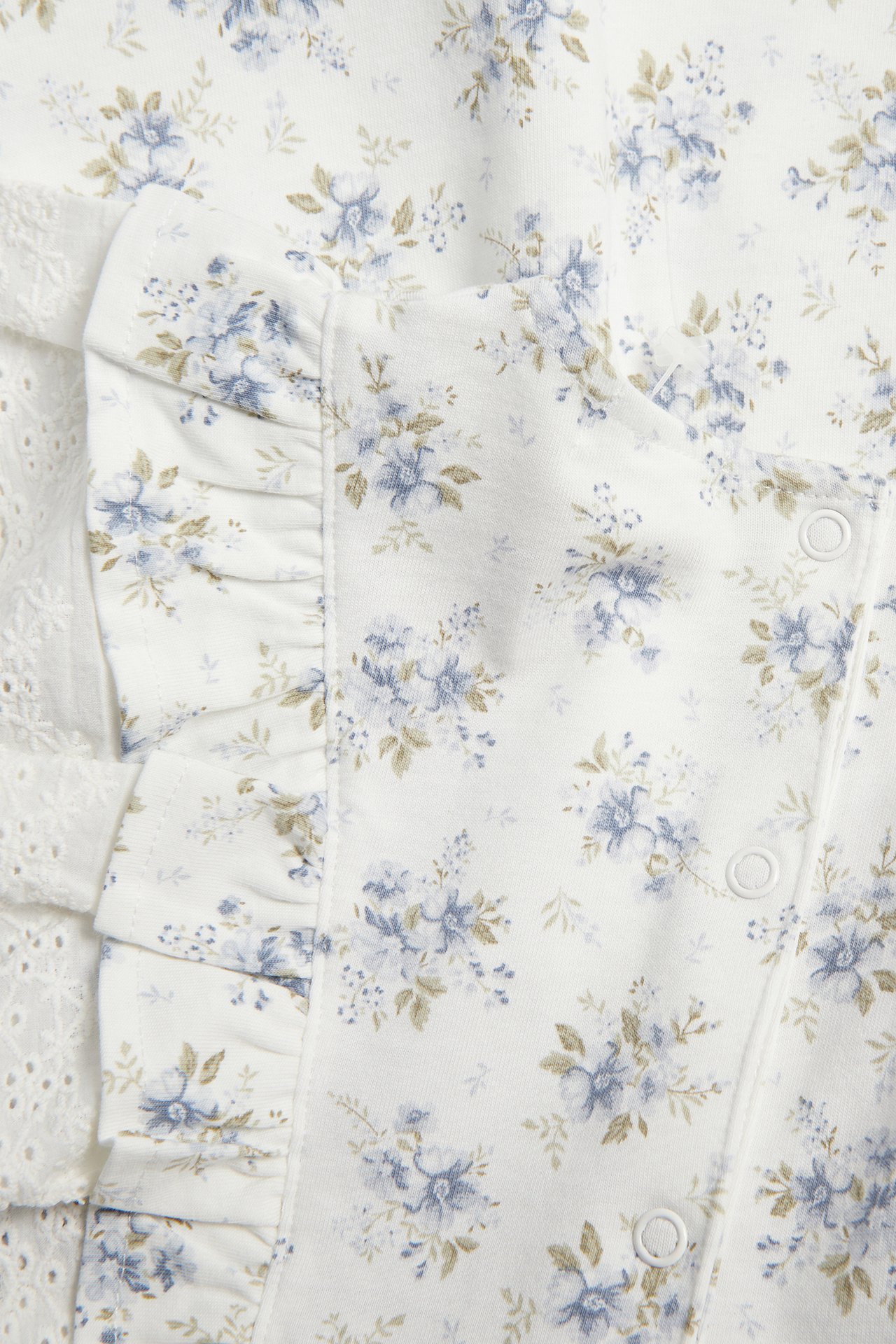 Nærbilde av en offwhite babykjole i blomstret jersey med blå blomster, volanger, hvit blonde og trykknapper.