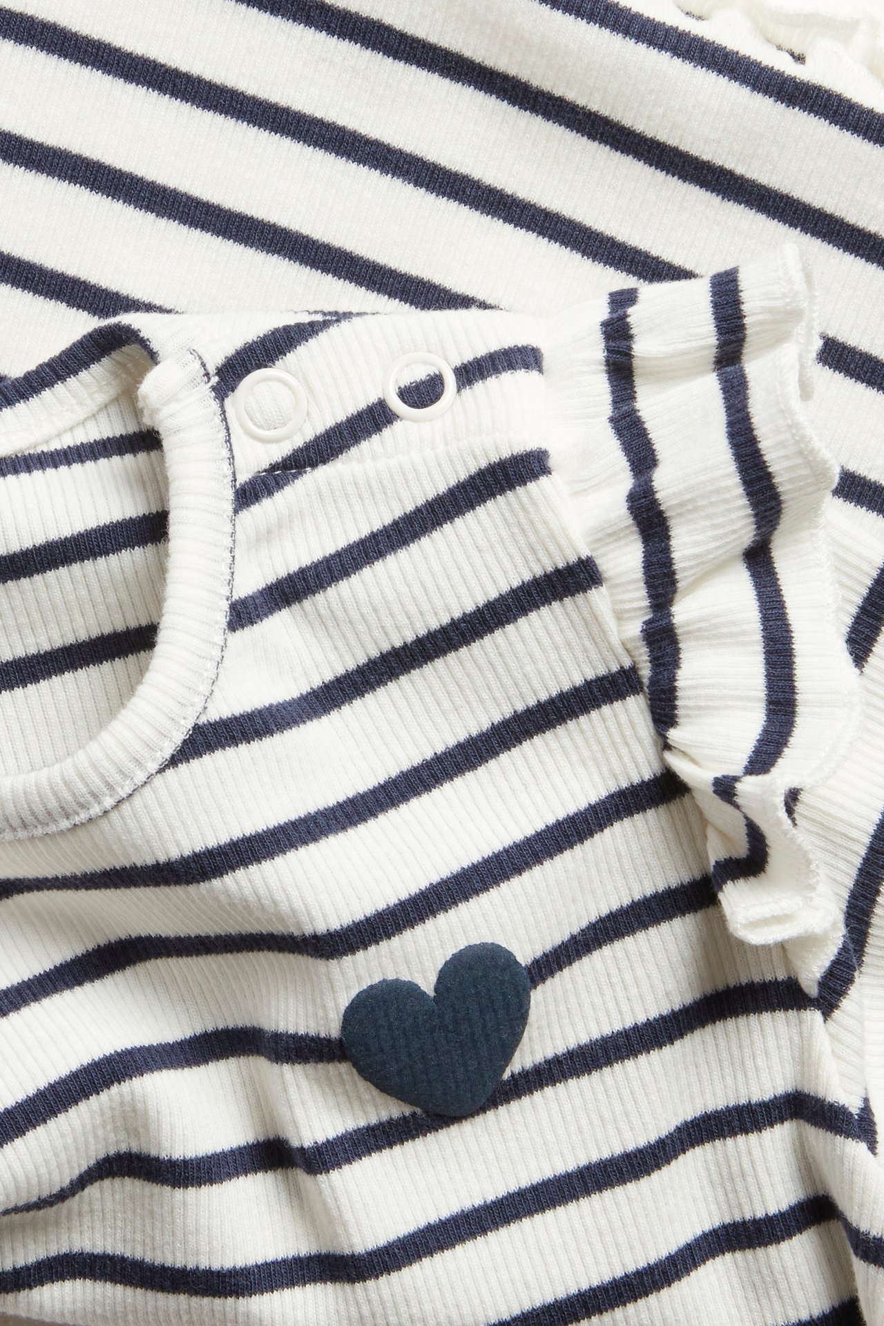 Nærbilde av babyens ribbestrikkede, offwhite- og blåstripede langermede topp med volanger og blått puffhjerte.