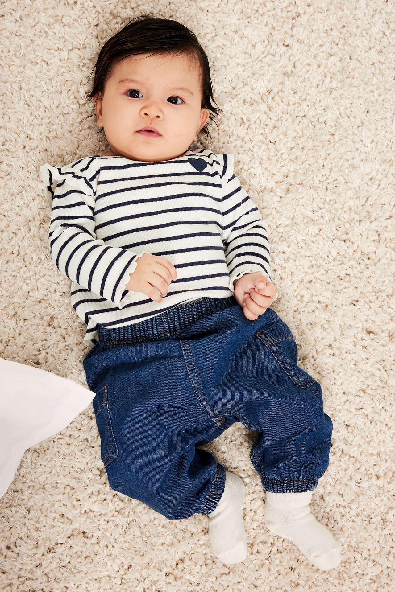 Oversiktsbilde av baby i hvit-blåstripet ribbet langermet topp med hjerte, blå bukser og hvite sokker.