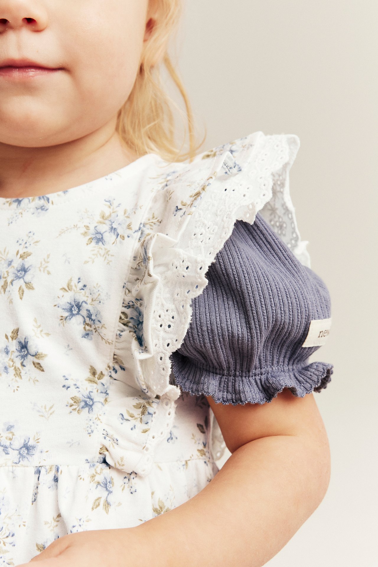 Lähikuva lapsesta yllään luonnonvalkoinen kukkakuvioinen trikooklänning pitsiröyhelöillä, alla ribattu violetinharmaa toppi.