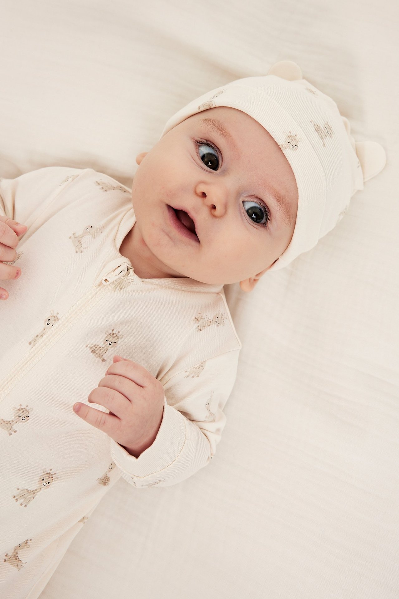 Nærbilde av baby i offwhite mønstret sparkedress med glidelås og matchende lue med ører.