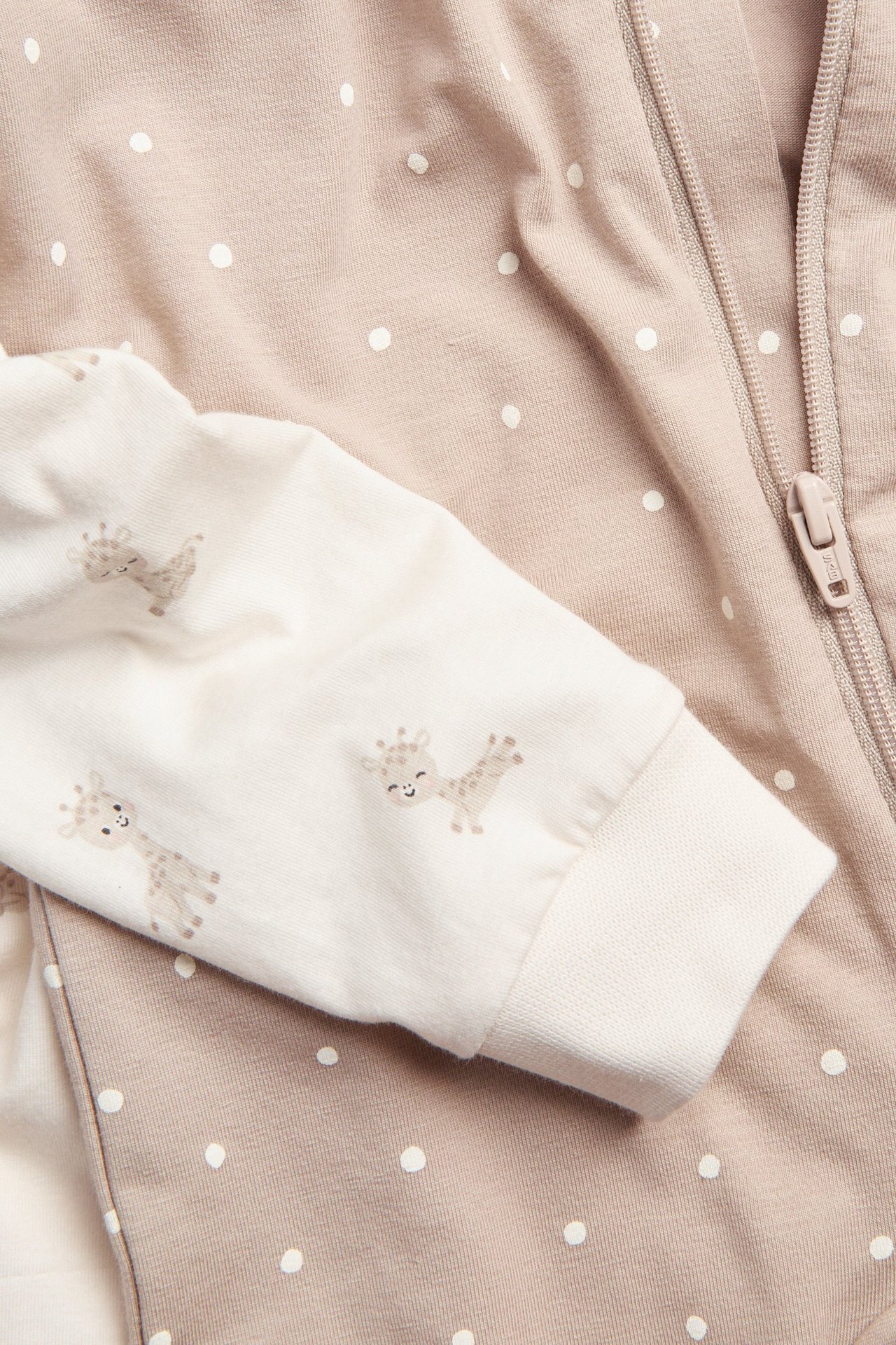 Lähikuva: Vauvan luonnonvalkoinen kirahvikuosinen potkupuvun hiha, beige pilkullinen potkupuku vetoketjulla.