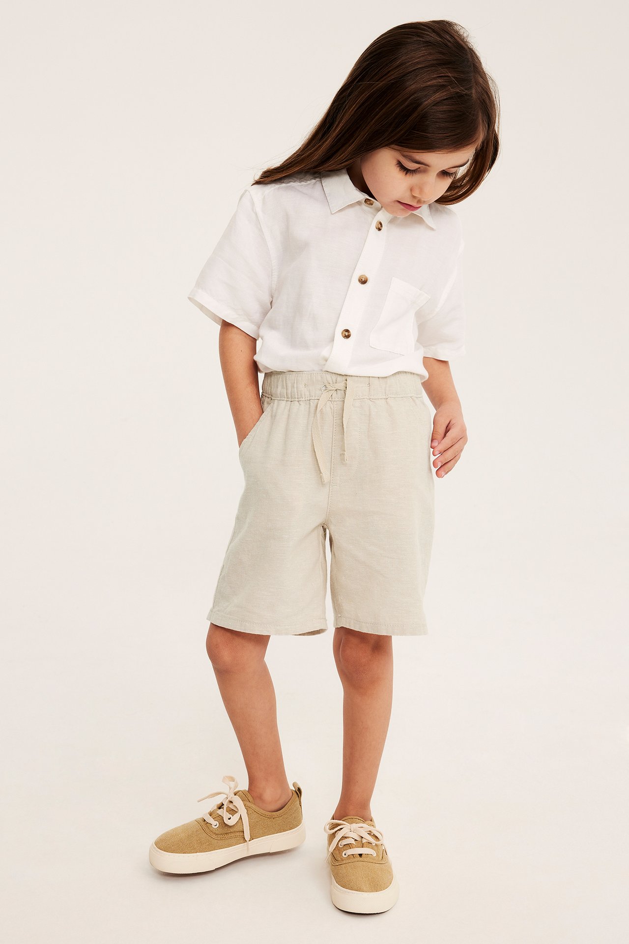 Barn i ljusbeige linneshorts med dragsko, vit kortärmad skjorta och ljusbruna sneakers.