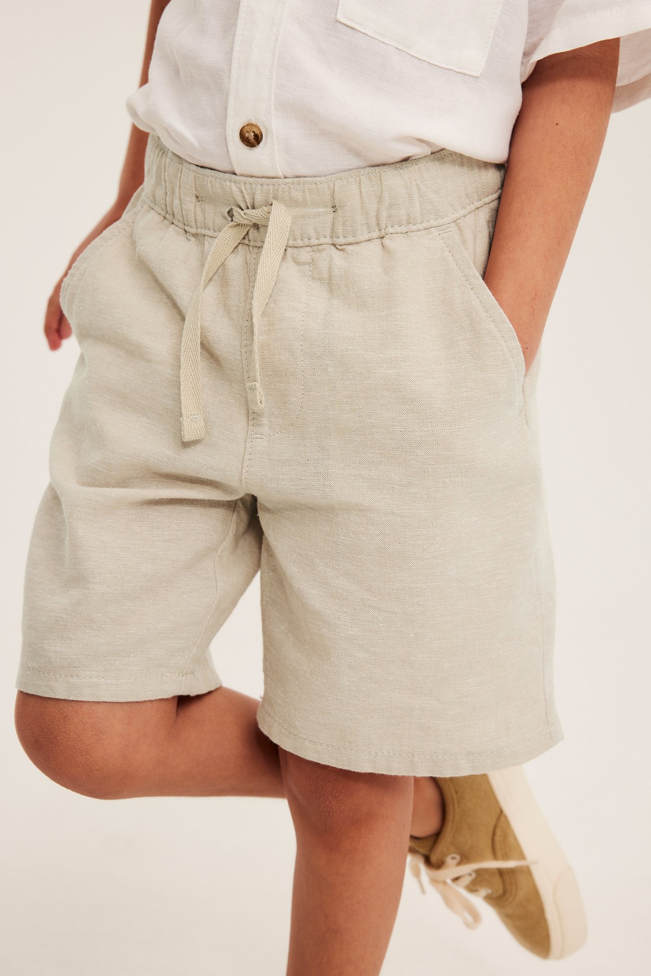 Frontvy: Barn bærer lys beige linshorts med snøring, hvit skjorte og oliven sneakers.