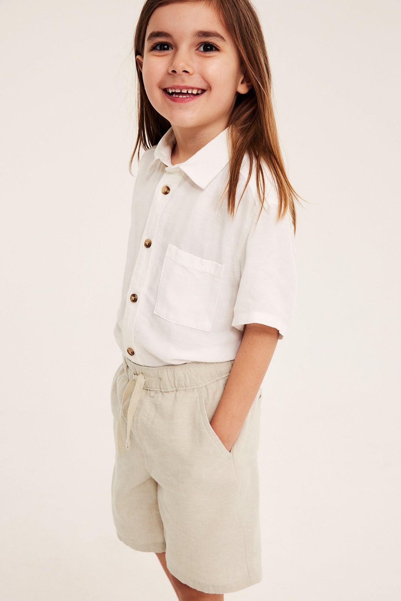Uśmiechnięte dziecko w jasnobeżowych lnianych szortach z troczkami i białej koszuli z krótkim rękawem.