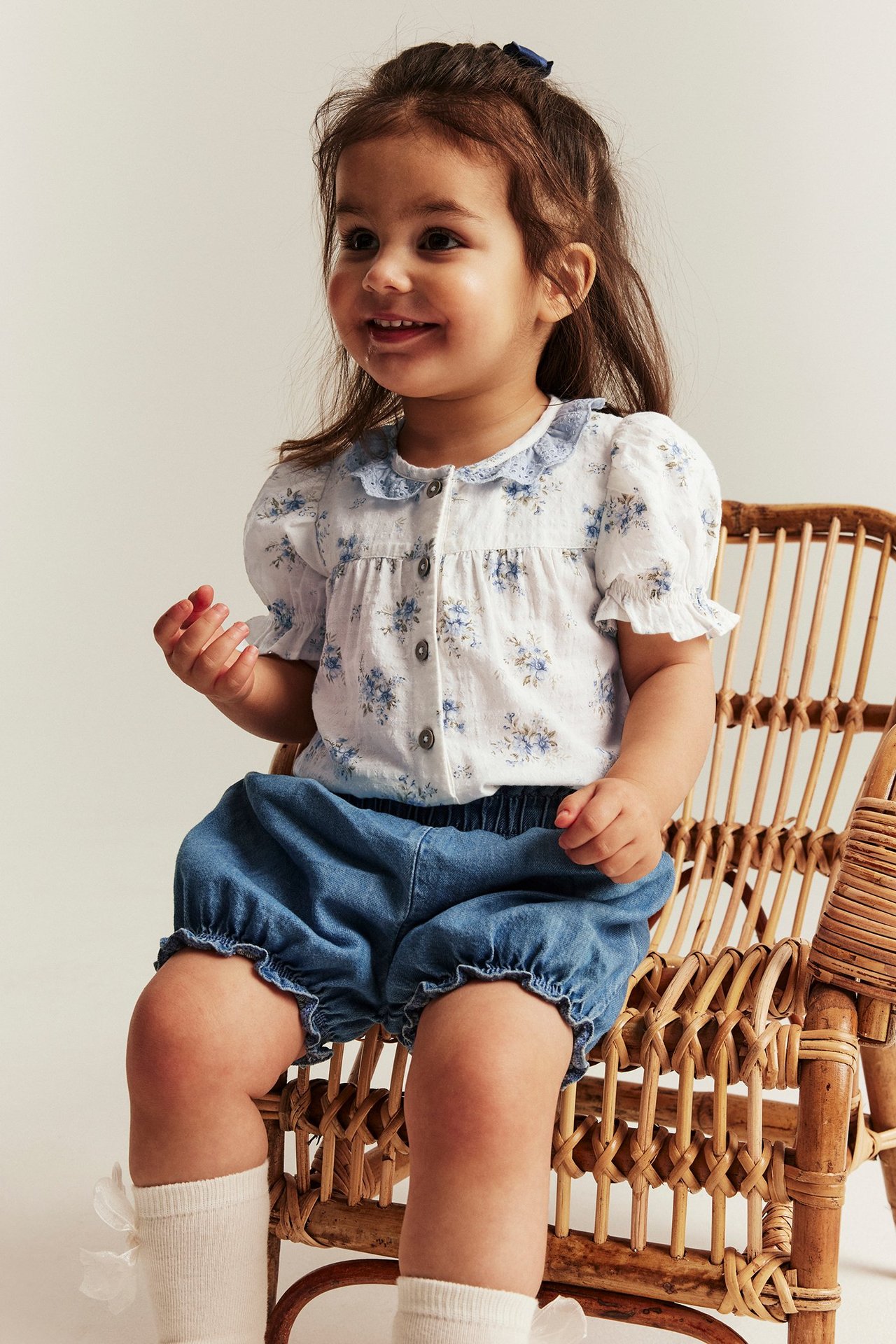 Frontbilde av en babyjente i offwhite blomstret kortermet body, blå shorts og hvite sokker.