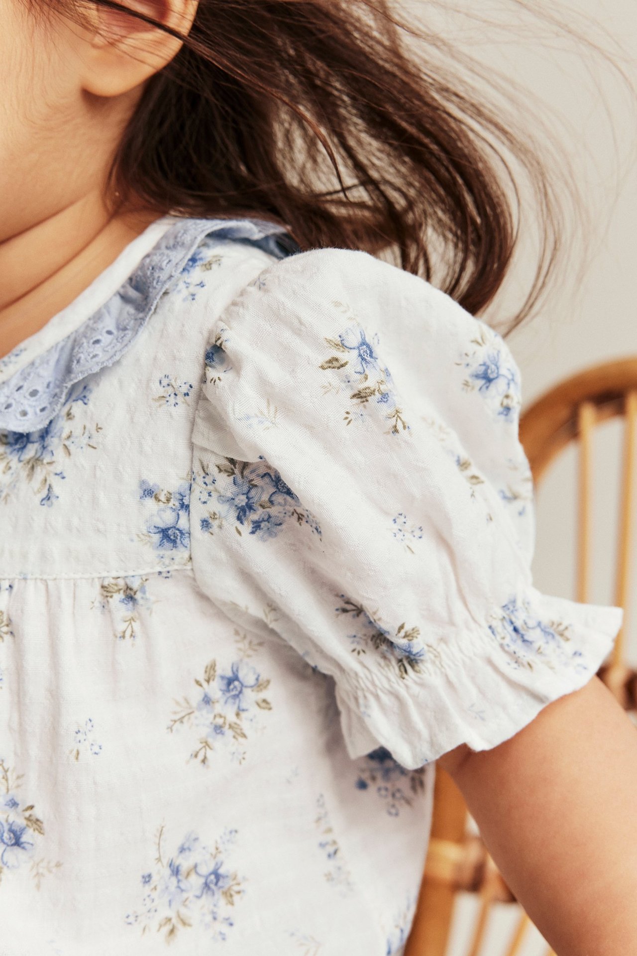 Zbliżenie: Dziecko nosi kremowy bawełniany body w kwiaty z niebieską koronkową falbaną i bufiastymi rękawami.