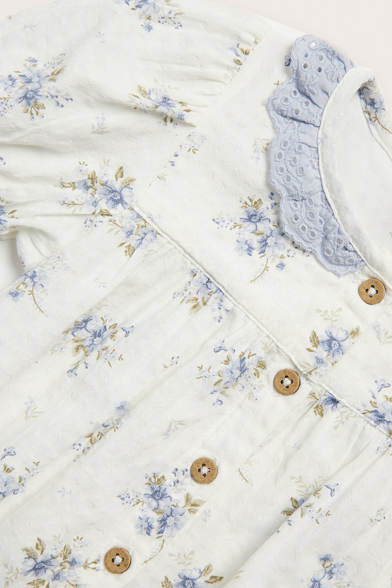 Zbliżenie na ecru body niemowlęce, niebieskie kwiaty, jasnoniebieski koronkowy kołnierzyk, guziki.
