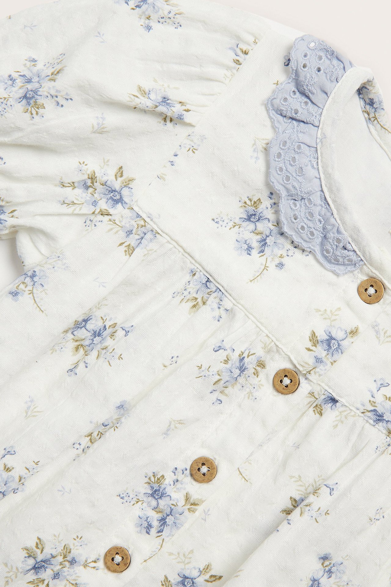 Zbliżenie na ecru body niemowlęce, niebieskie kwiaty, jasnoniebieski koronkowy kołnierzyk, guziki.