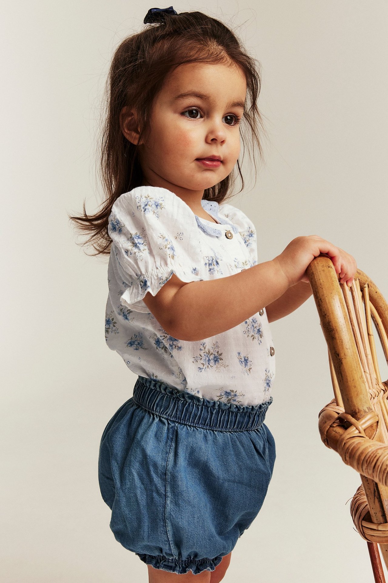 Dziewczynka z boku w kremowym kwiecistym body, niebieskich szortach, trzyma krzesło rattanowe.