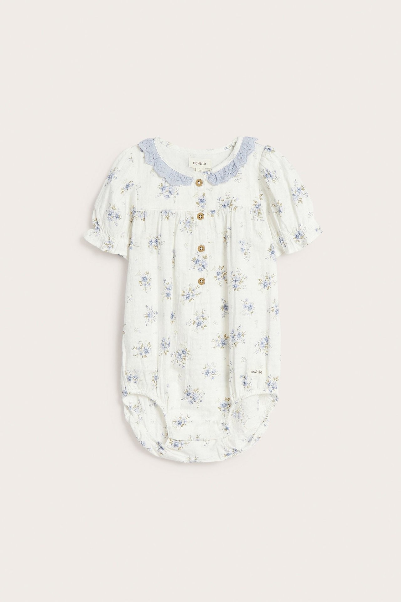 Frontvisning av en offwhite babybody med blått blomstermønster, lyseblå blondekrage og korte puffermer.