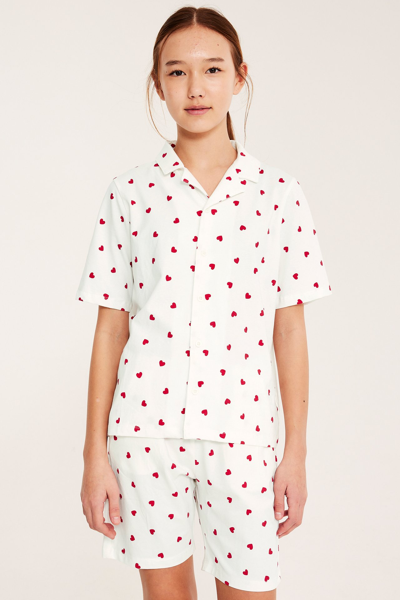 Frontbilde av barn i en off-white bomullspyjamas med rødt hjertemønster, kortermet skjorte og shorts.