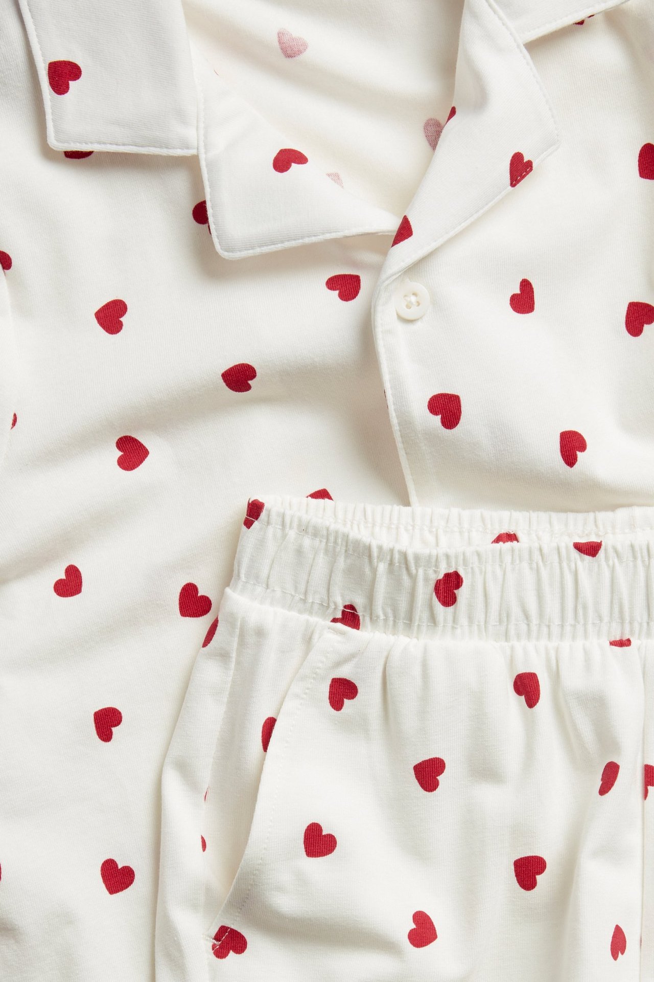 Nærbilde av offwhite pysjamas i bomullsblanding med rødt hjertemønster, med kortermet skjortetopp og shorts med strikk.