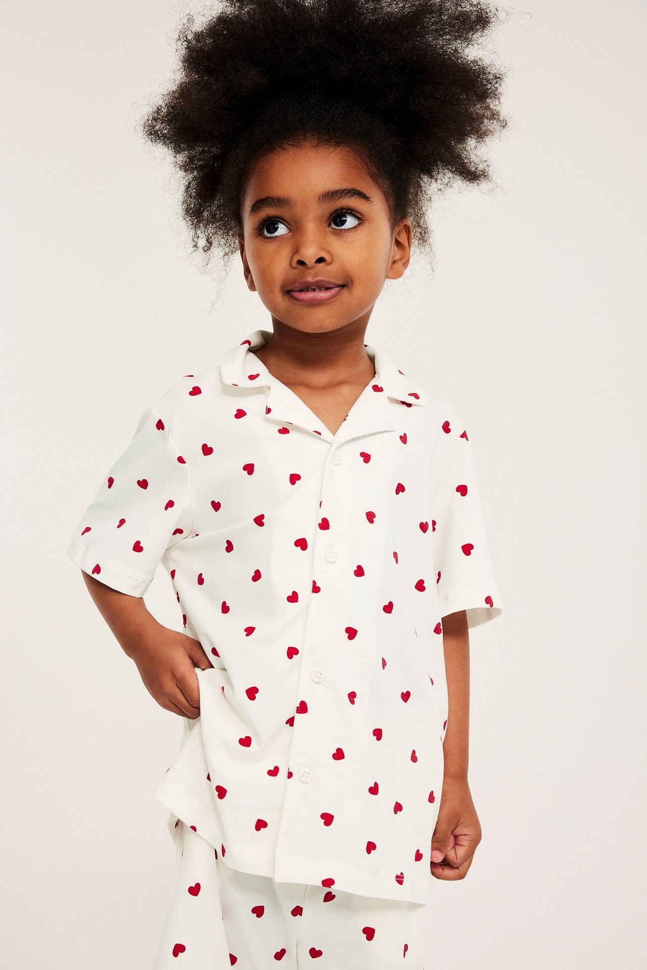 Forfra: Et barn i en offwhite, hjertemønstret pyjamas med kortermet skjorte og bukser.