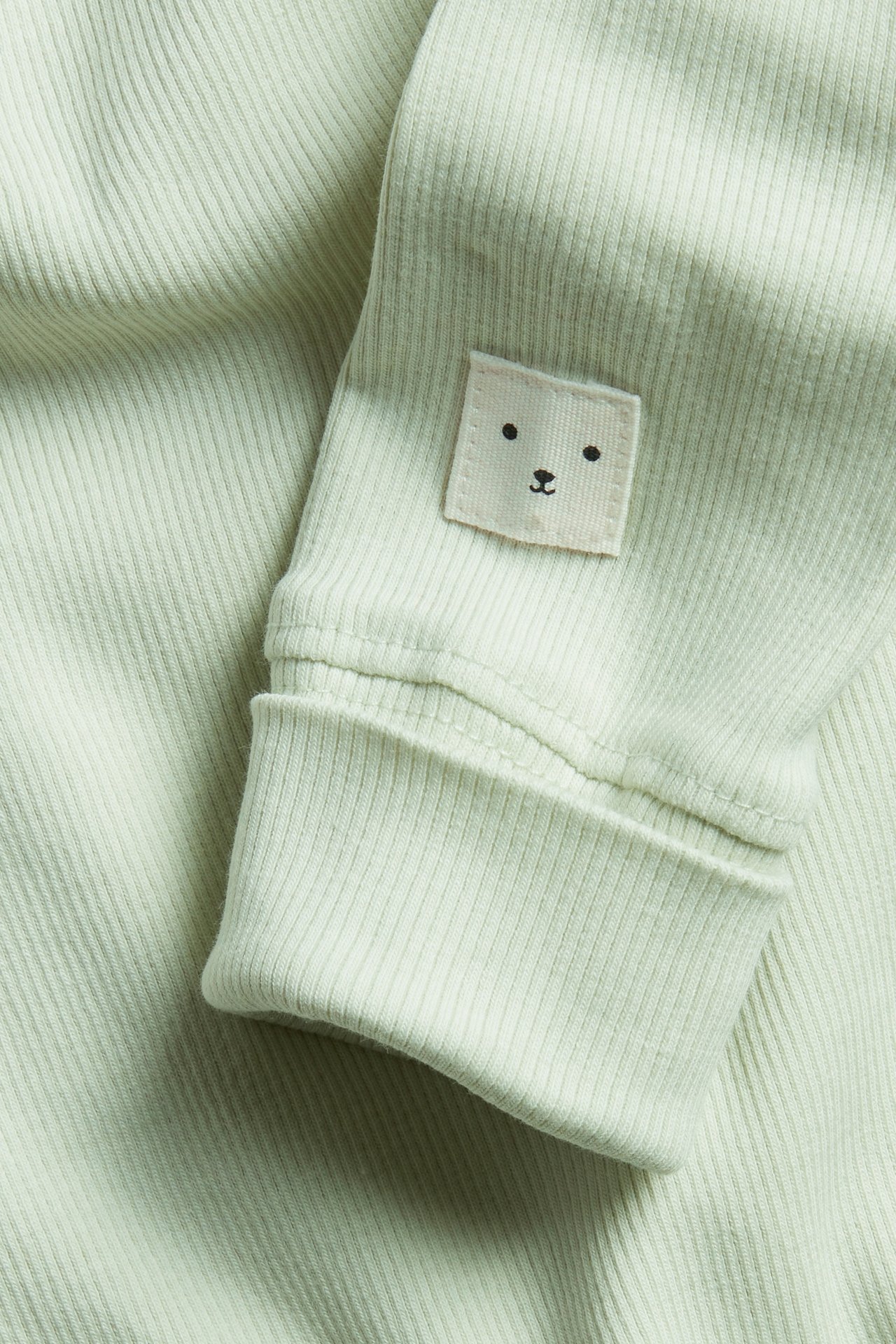 Zbliżenie rękawa jasnozielonego prążkowanego body niemowlęcego z naszywką misia i podwiniętym mankietem.