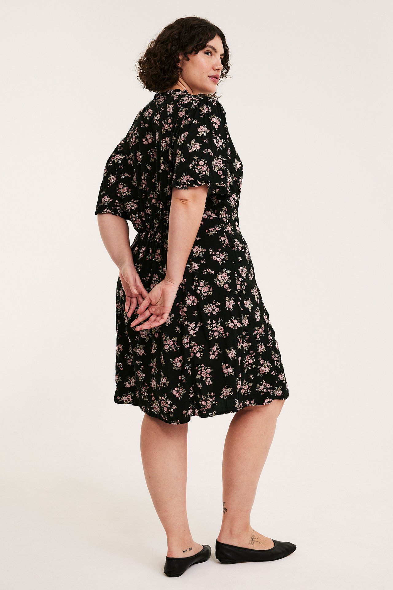 Sidovy: Kvinna i svart knälång blommig viskosklänning med volanghals, korta ärmar och svarta ballerinaskor.
