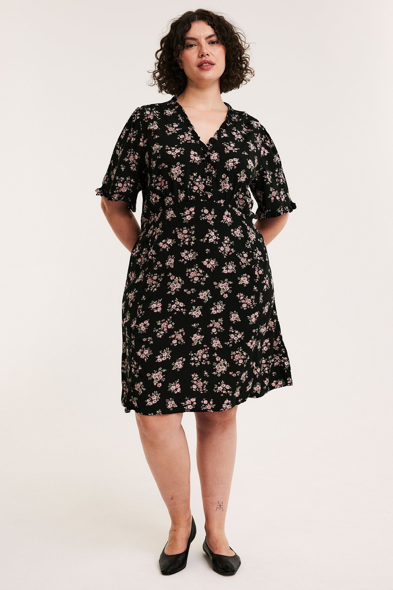 Etunäkymä: Nainen mustassa kukkakuosisessa V-kaulaisessa polvipituisessa mekossa röyhelöillä ja napeilla, mustat kengät.