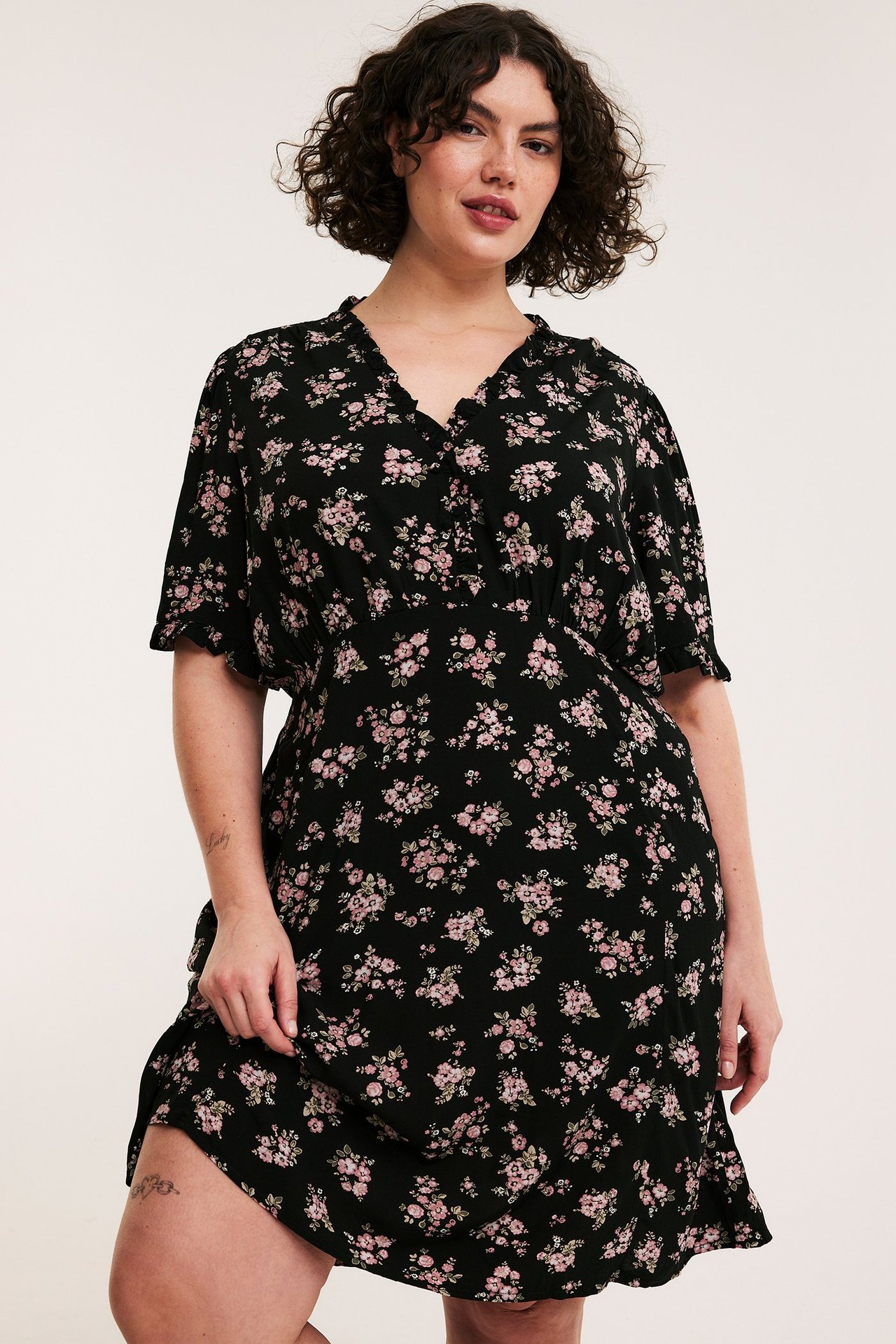 Edestäpäin, nainen mustassa kukkakuosisessa polvipituisessa mekossa, jossa röyhelökaulus ja lyhyet hihat.