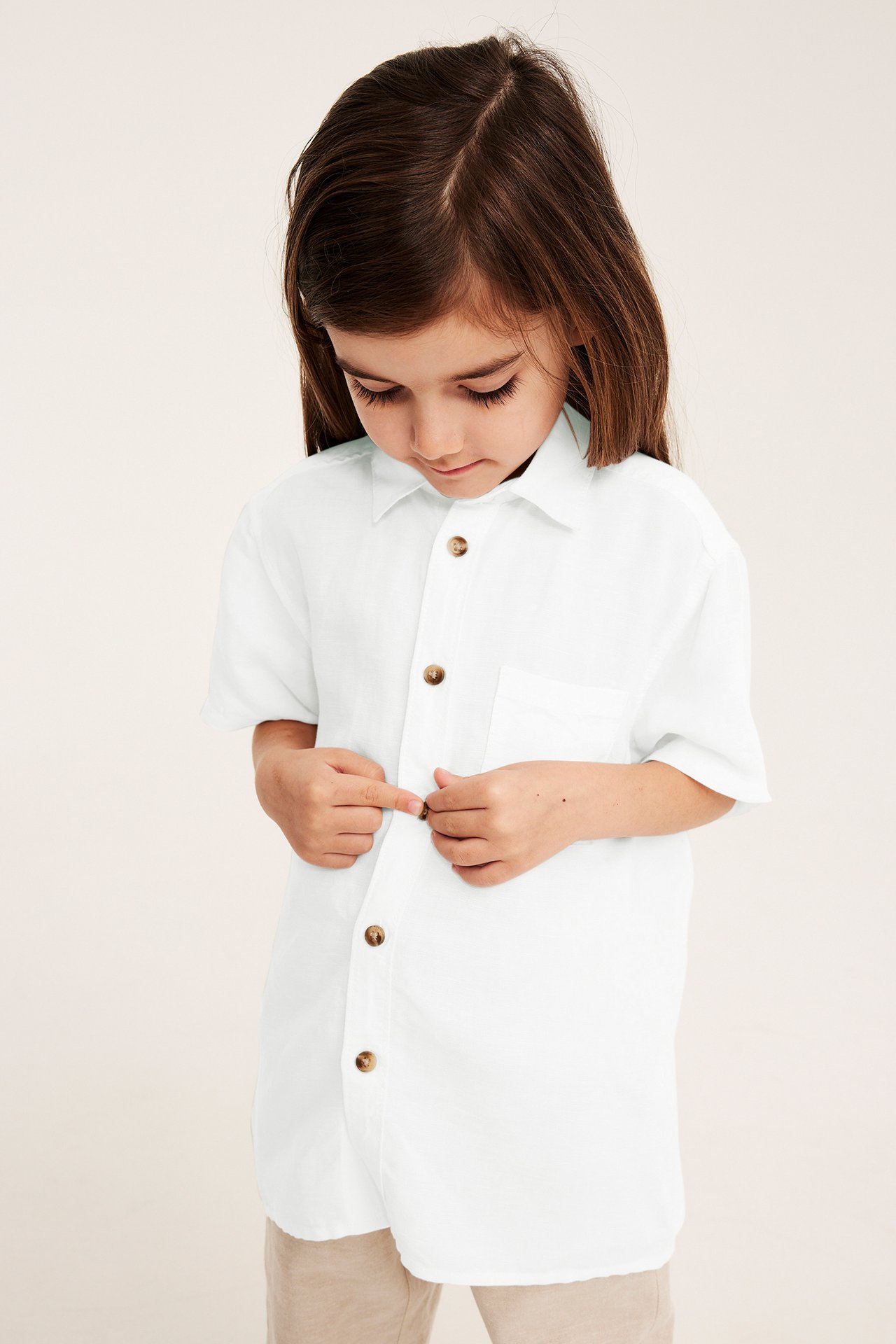 Frontvy av ett barn som knäpper en vit kortärmad skjorta i linnemix med bruna knappar och beiga byxor.