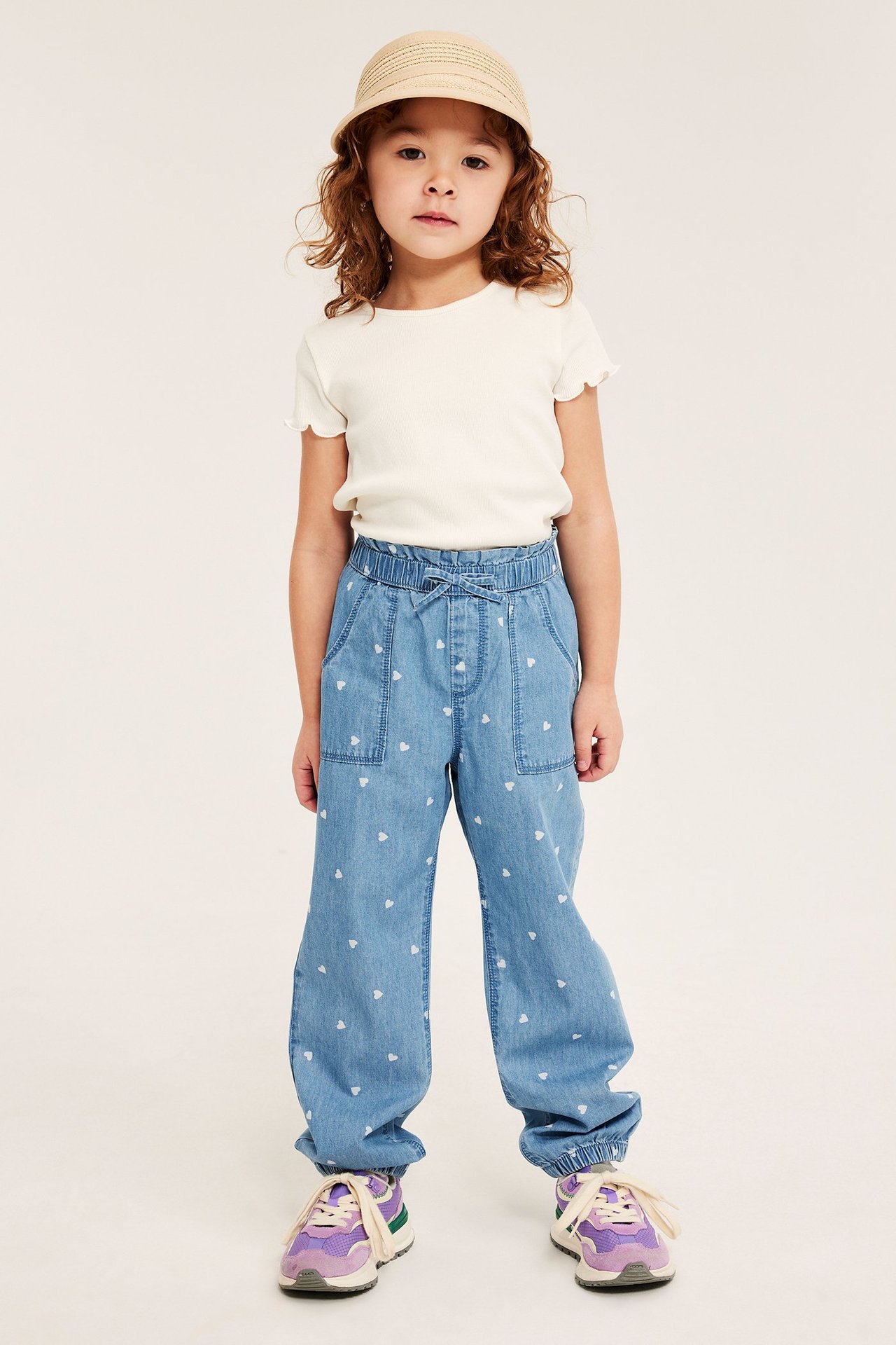 Framifrån: Barn i ljusblå hjärtmönstrade denimbylxor, gräddvit topp, stråhatt och lila sneakers.