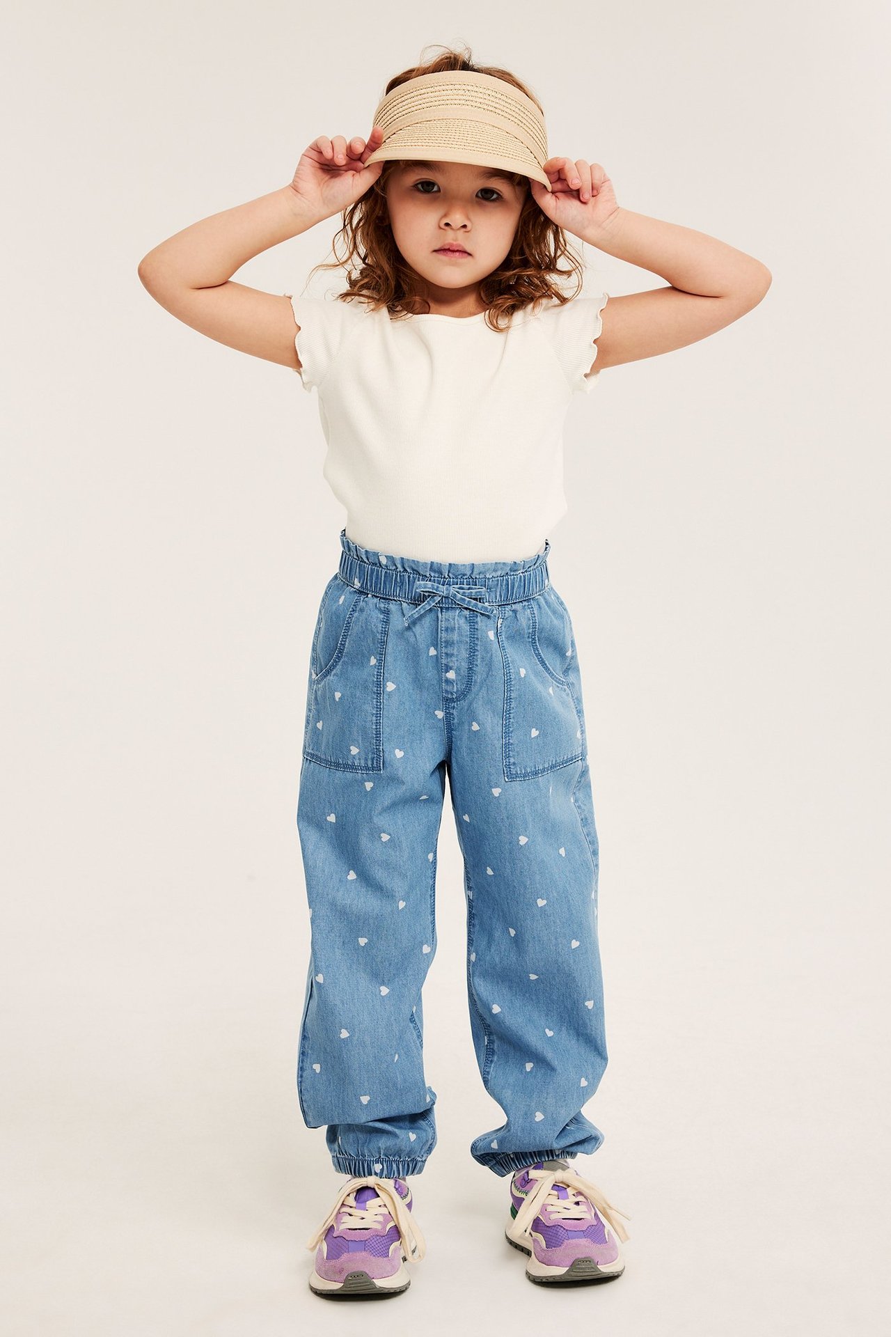Barn bär ljusa denimbyxor med hjärtmönster, vit t-shirt, beigt solvisir och lila sneakers. Frontvy.