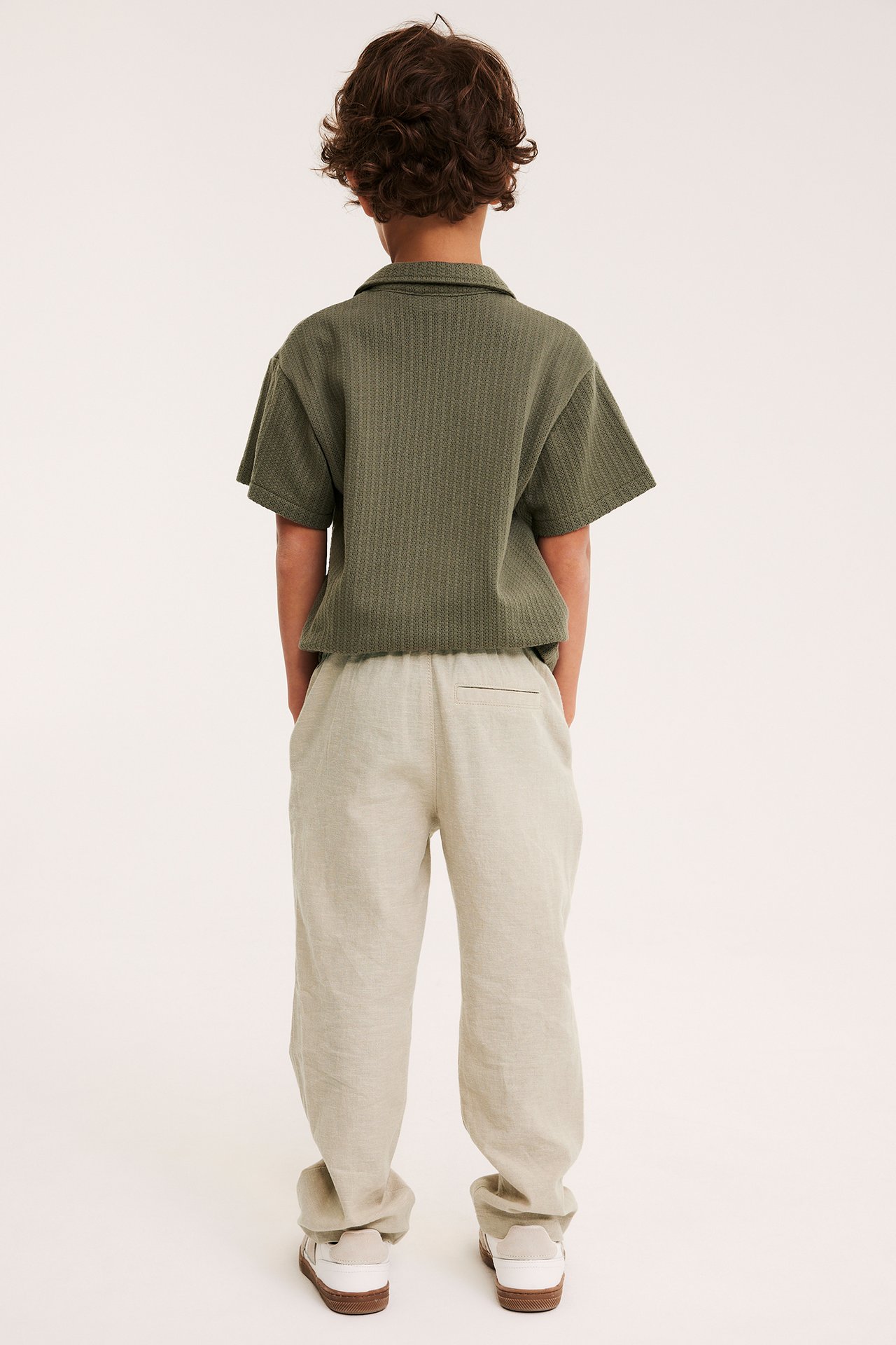 Bakfra: Et barn i beige linblandingsbukser, en grønn strukturert skjorte og hvite joggesko.