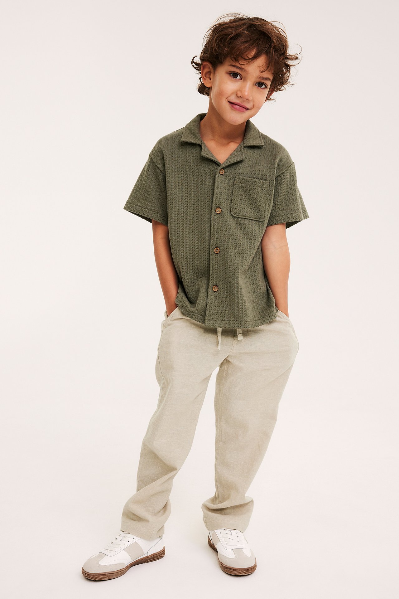 Frontbild på ett barn i beiga dra-på-byxor i linnemix, grön kortärmad skjorta och vita sneakers.