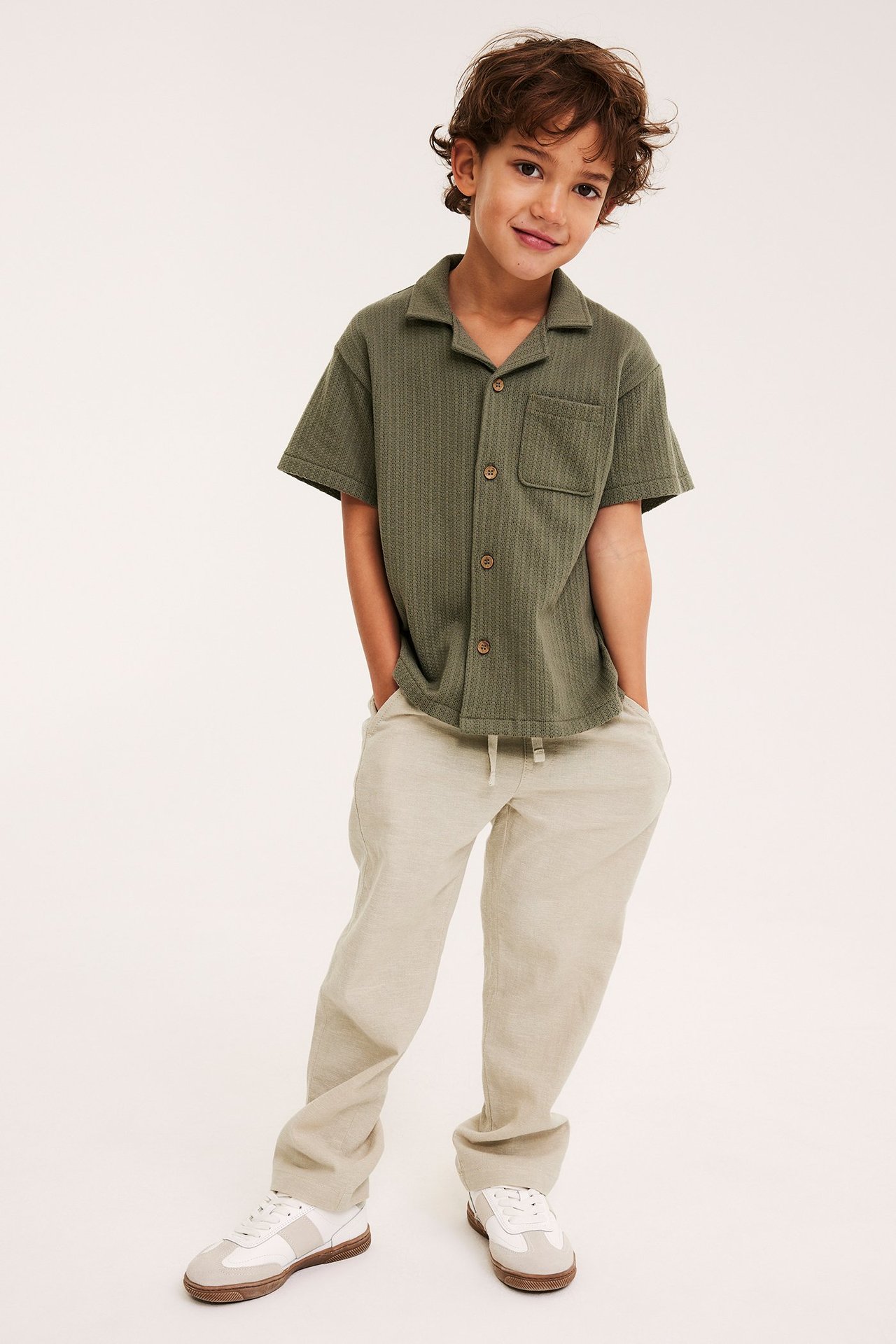 Edestä: lapsi beigessä pellavasekoitehousuissa, vihreässä lyhythihaisessa paidassa ja valkoisissa lenkkareissa.