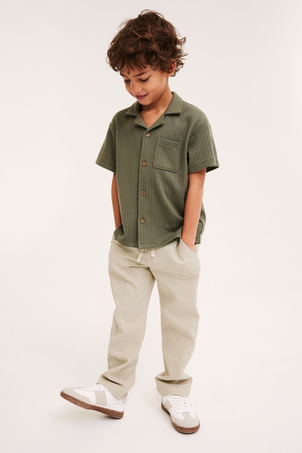 Edestä kuvattu lapsi beigessä pellavasekoitevetohousuissa, vihreässä tekstuuripaidassa ja valkoisissa tennareissa.