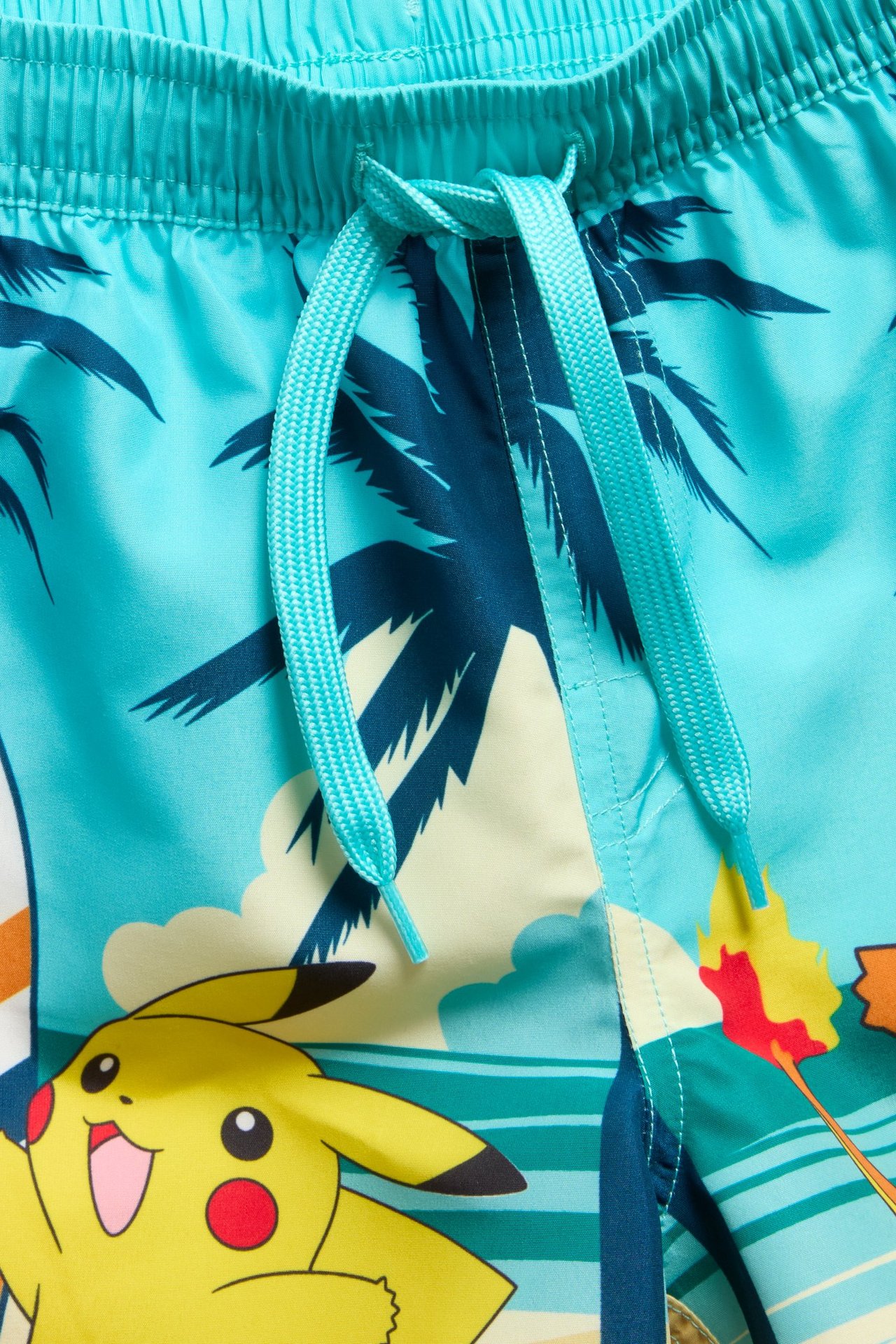 Lähikuva, edestä, lasten vaaleansinisistä uimahousuista, joissa kiristysnyöri ja värikäs Pokémon-kuvio.