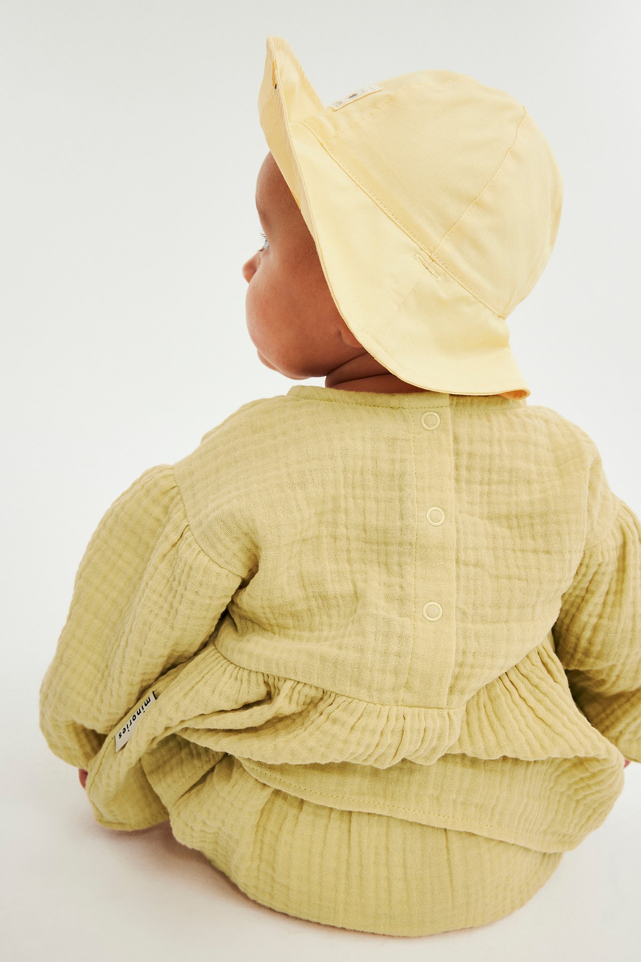Bakifrån på en baby i en ljusgul muslinklänning, matchande bloomers och en gul solhatt.