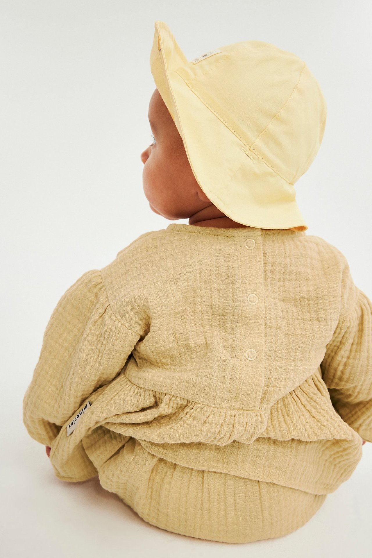 Bakifrån: baby i en beige muslinklänning, matchande bloomer och en ljusgul solhatt.