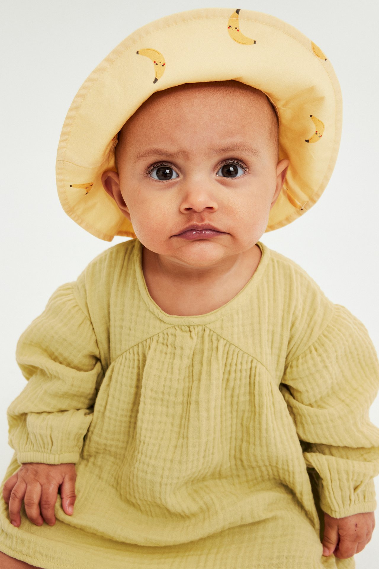 Frontvy, baby i gulgrön musslinklänning med rynkad överdel och ljusgul hatt med bananmönster.