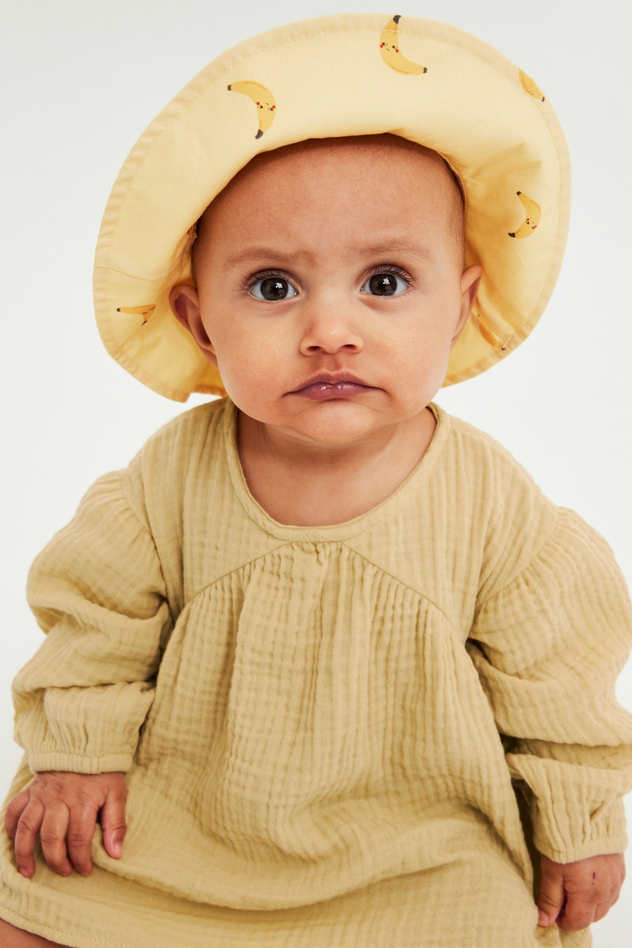 Edestäpäin: vauva vaaleankeltaisessa musliinimekossa rypytetyllä kaarrokkeella ja keltaisessa banaanikuvioisessa lierihatussa.