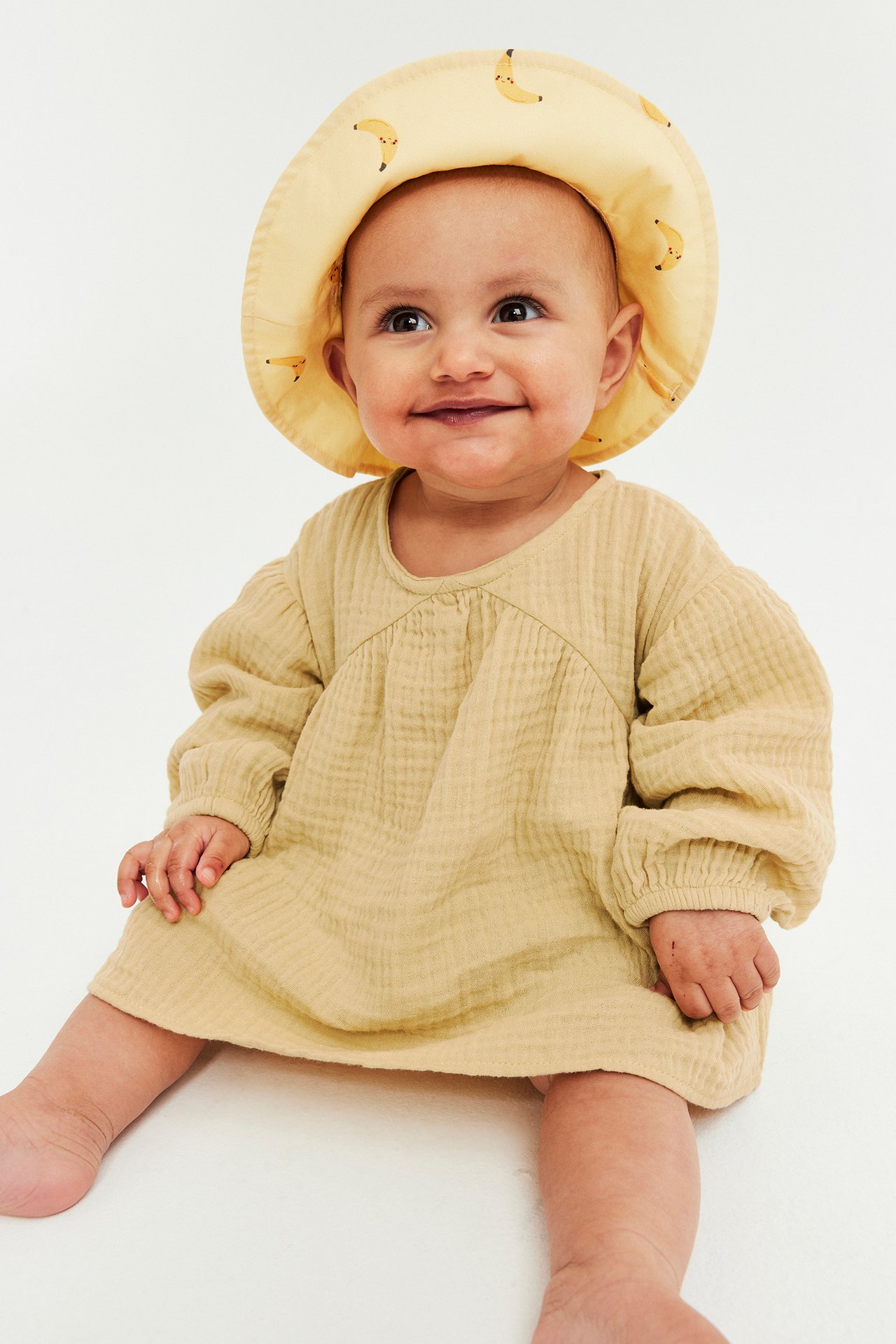 Edestä kuvattu hymyilevä vauva yllään löysä keltainen musliinimekko ja keltainen banaanihattu.