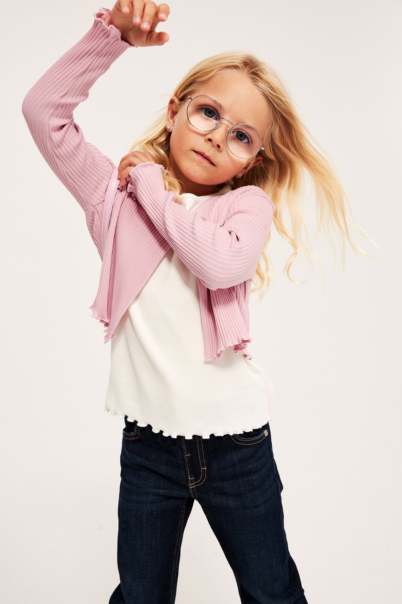 Frontbilde av et barn i lys rosa ribbet kort bolero over hvit topp, mørkeblå jeans og runde briller.