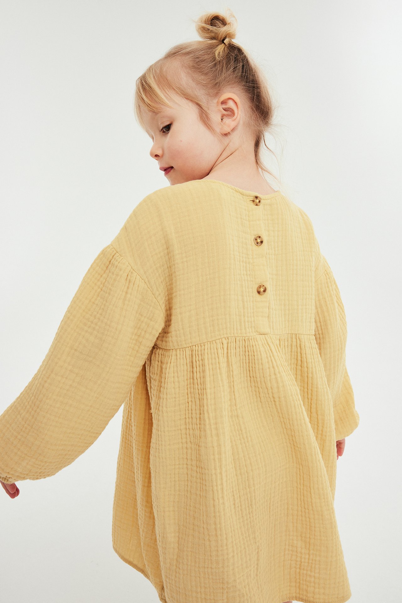 Widok z tyłu dziecka w luźnej, żółtej sukience muślinowej z długimi rękawami i trzema brązowymi guzikami.