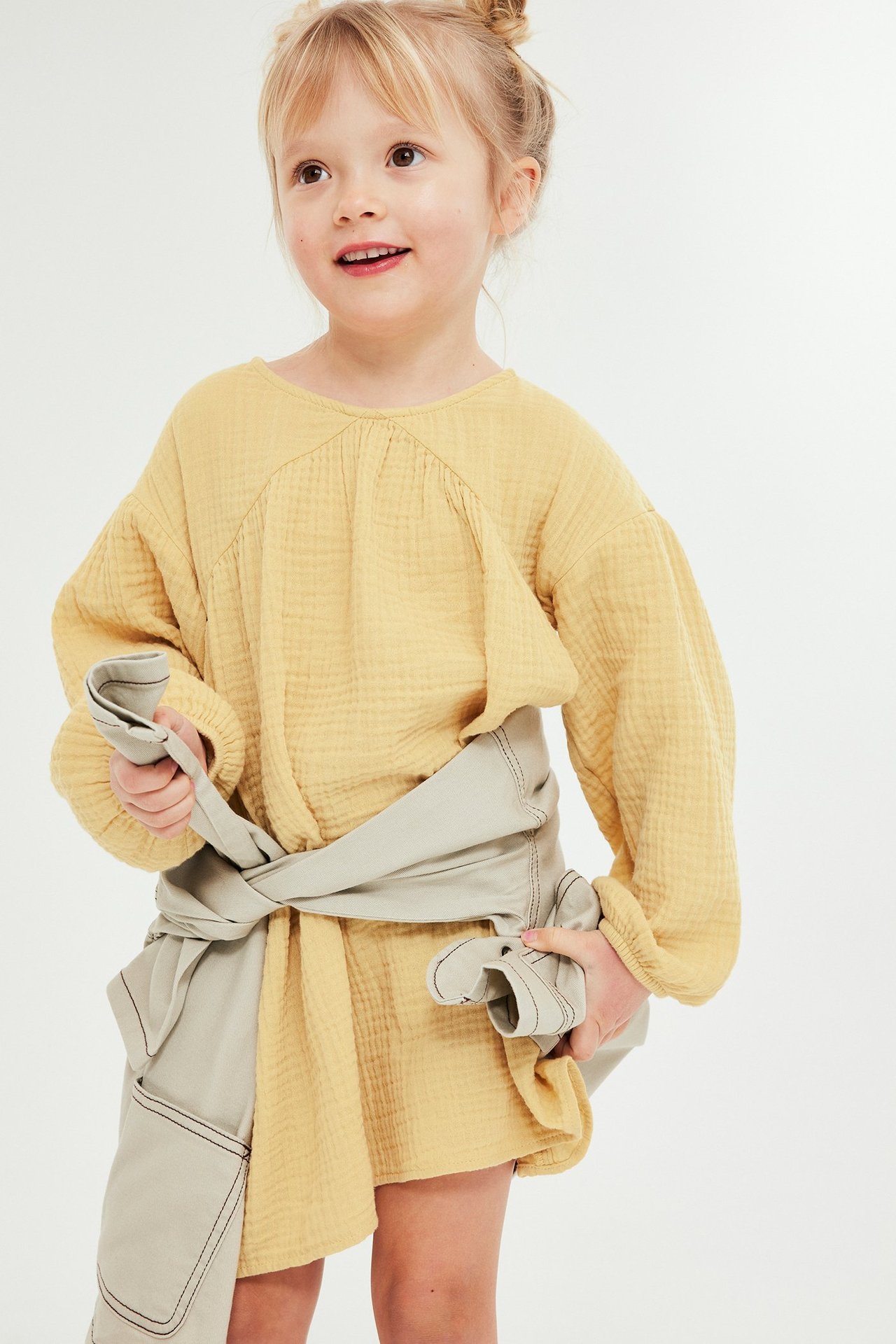 Frontvisning av et barn i en løs gul musselin-kjole med et lysbeige plagg knyttet rundt midjen.