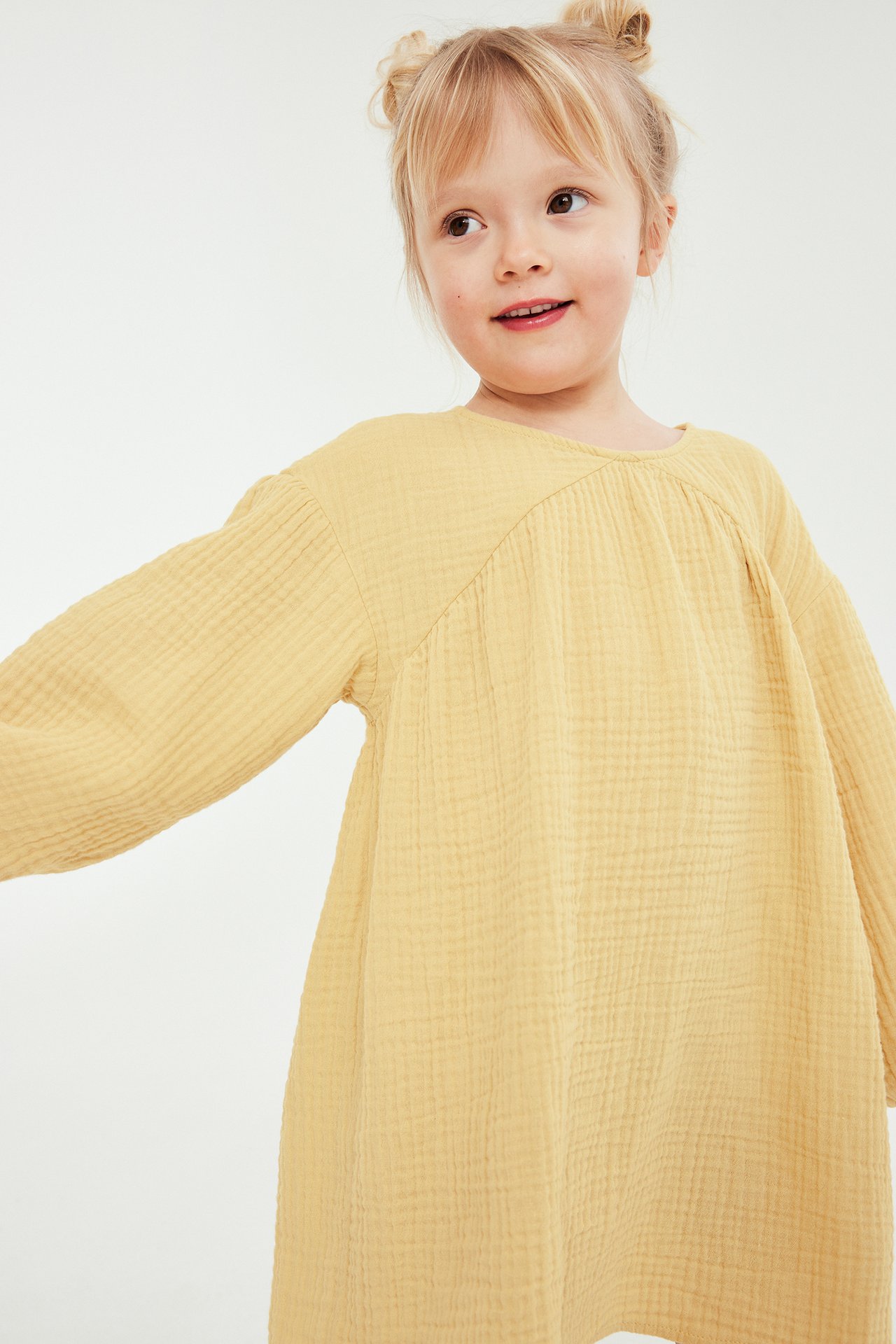 Dziecko w luźnej żółtej sukience z bawełnianego muślinu, z długimi rękawami i marszczonym dekoltem.